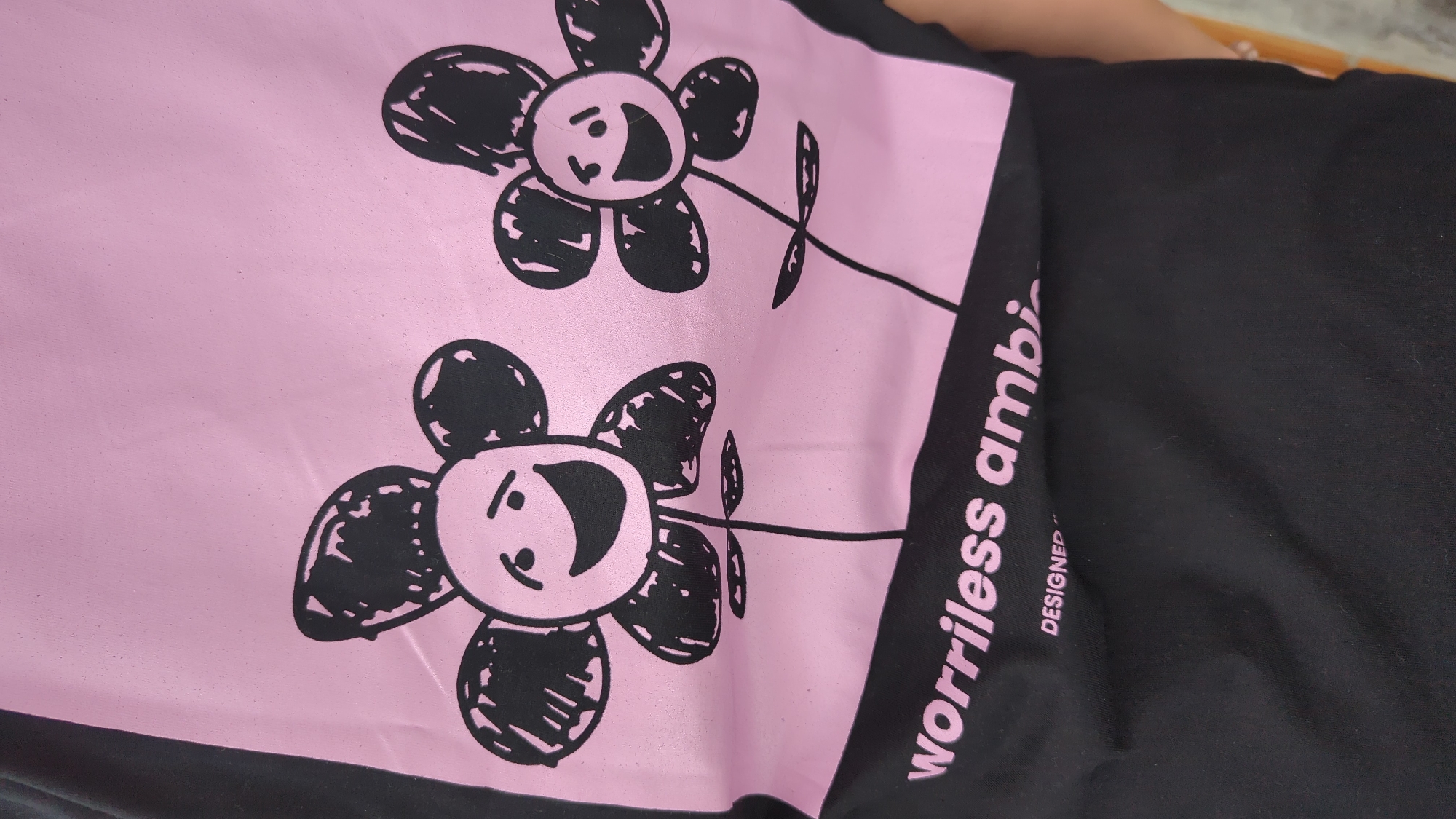 A close-up shot shows the detail of a black t-shirt with a pink graphic. The graphic features two cartoon-like flowers with faces drawn in a child-like, scribbled style. The flowers have exaggerated smiles and beady eyes. Below the flowers, text reads "worriless ambi" in a playful, handwritten font. Underneath this, the word "DESIGNED" is printed in smaller text. The t-shirt is black and appears to be made of a soft fabric. The image is taken from a slightly elevated angle, focusing on the print and the texture of the material. The background is out of focus and suggests an indoor or outdoor setting, possibly in North Kuta, Indonesia, though no specific landmarks or identifying features of the location are visible.