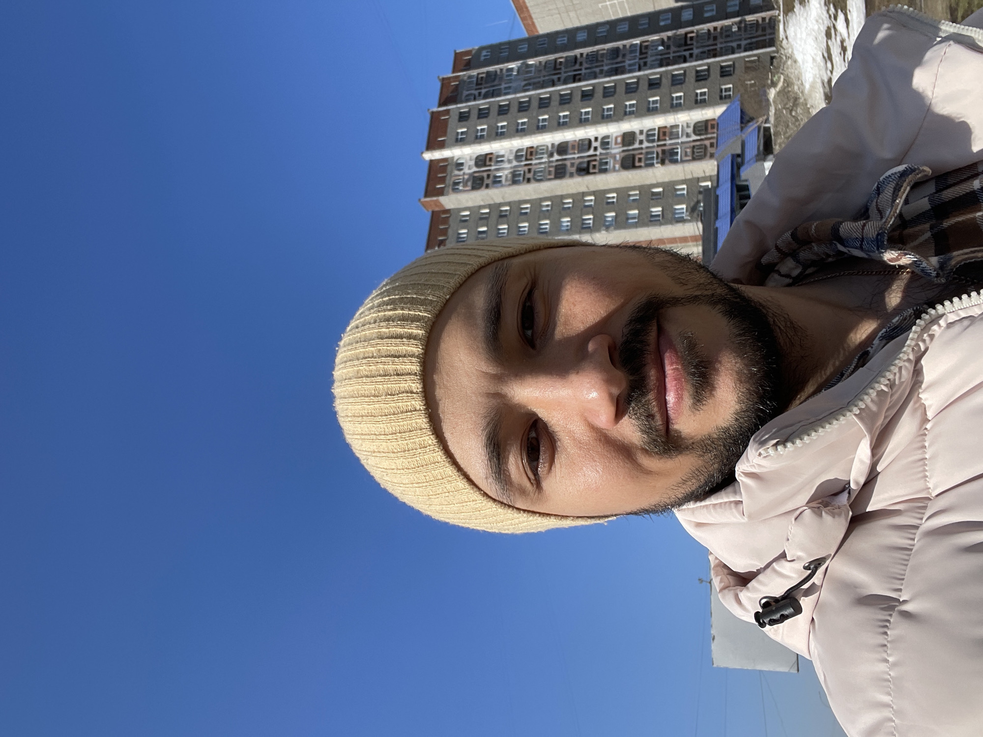 A man with dark hair and a beard is wearing a beige knit beanie and a light pink puffer jacket with a plaid shirt underneath. He is standing outside in front of a tall, rectangular building with rows of windows and a clear blue sky. The building is likely located in Izhevsk, Russia.