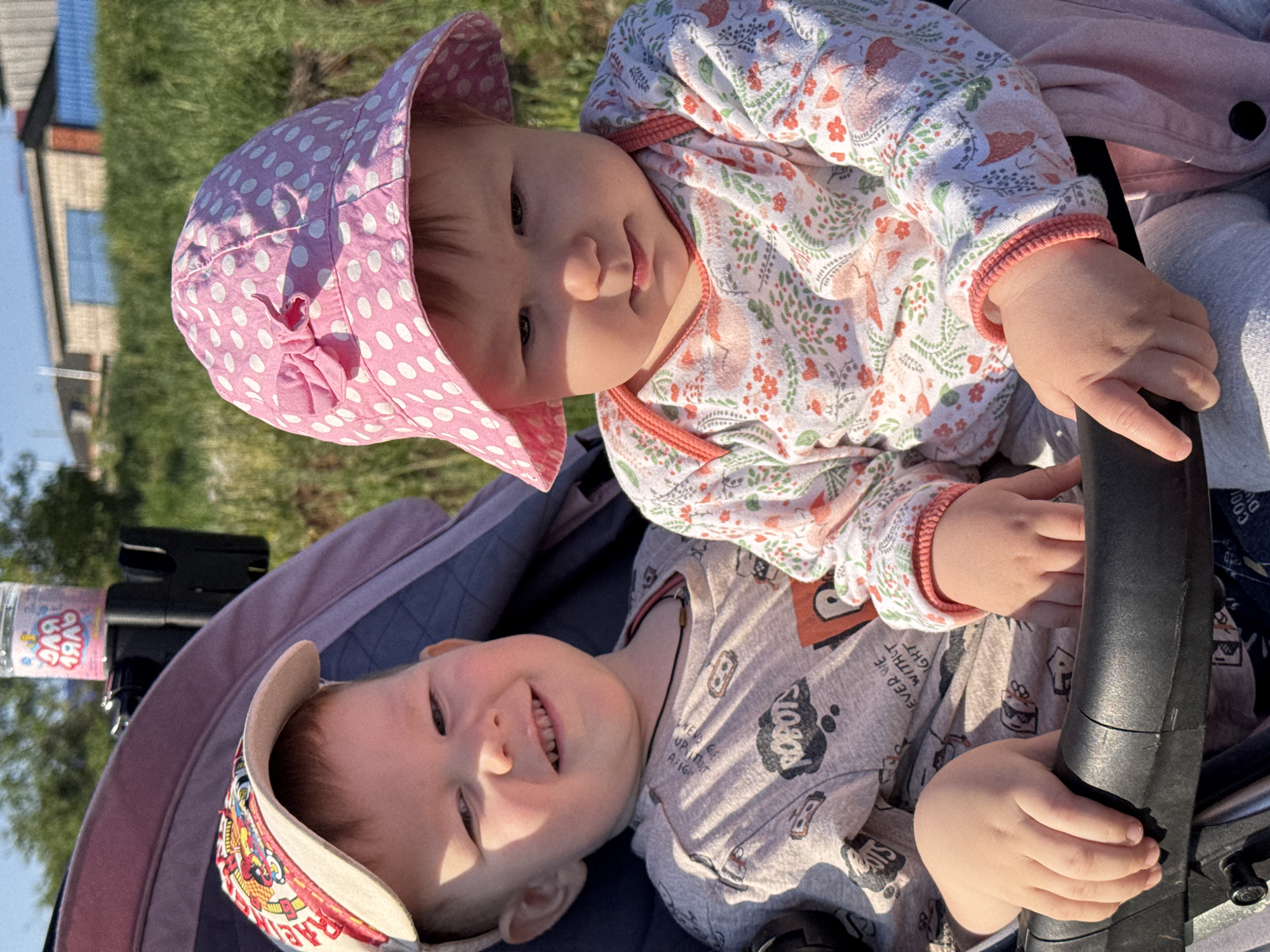 Two young children are in a stroller, with the photograph taken from above. The child on top wears a pink polka-dot sun hat and a floral-patterned top. The child below wears a baseball cap and a grey printed pajama shirt. They are both holding onto the handlebar of the stroller. A water bottle is visible on the left side of the stroller. The background shows a grassy area, a building, and a blue sky. The location might be Zav'yalovo, Russia, but there are no buildings visible that would confirm the location.