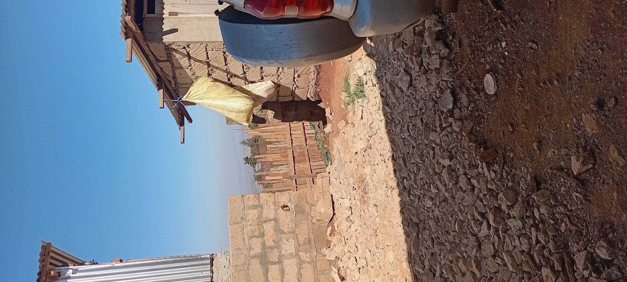 The image depicts an outdoor scene in Kijabe ward, Kenya, characterized by a rough, unpaved ground covered in stones and dirt. In the foreground, a shadow of what appears to be a vehicle is cast across the rocky surface. The middle ground reveals a fence constructed from wooden planks, partially overgrown with sparse green vegetation. Beyond the fence, a landscape of hills and distant structures is visible under a clear blue sky. The immediate area features construction elements, including a wall made of stacked rectangular stones. A large, yellow bag is hanging from a wooden beam above this wall, secured by a blue string. To the right, a portion of a silver vehicle, likely a car, is visible with its taillight showing. The overall impression is one of an unfinished construction site or a rural dwelling in an arid or semi-arid environment.