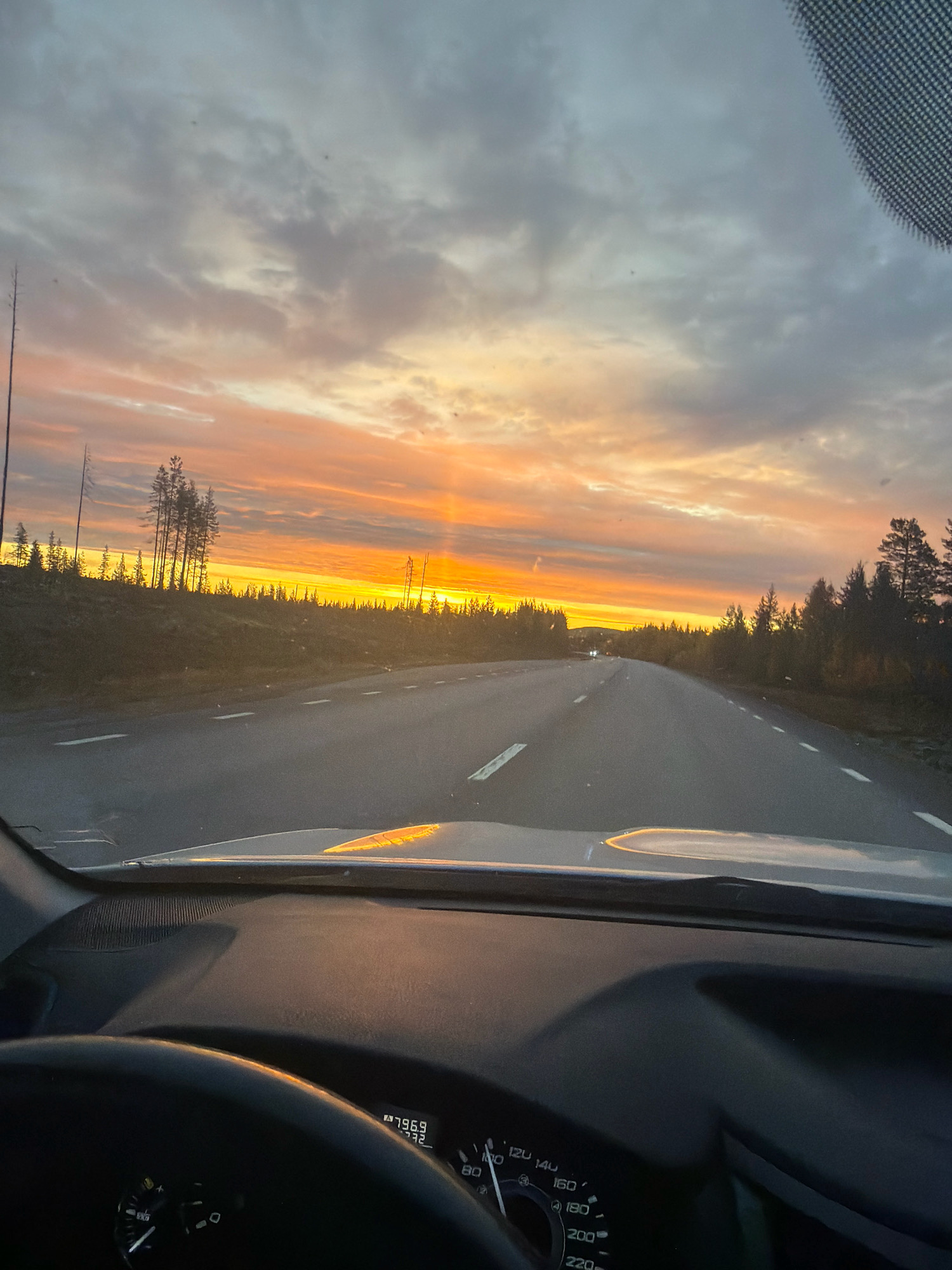 The image shows a view from inside a car driving on a road at sunset. The sun is low in the sky, casting a warm orange and yellow glow through the clouds, illuminating the road and the surrounding landscape of trees and hills. The road is paved with white markings and appears to be in a rural or wilderness area, possibly near Gällivare, Sweden, given the terrain and vegetation which are consistent with the subarctic climate and boreal forest characteristic of that region. The dashboard of the car is visible in the foreground, with a steering wheel and part of the instrument cluster showing speed and other readings. There are no people or other vehicles clearly visible on the road, suggesting a solitary journey.