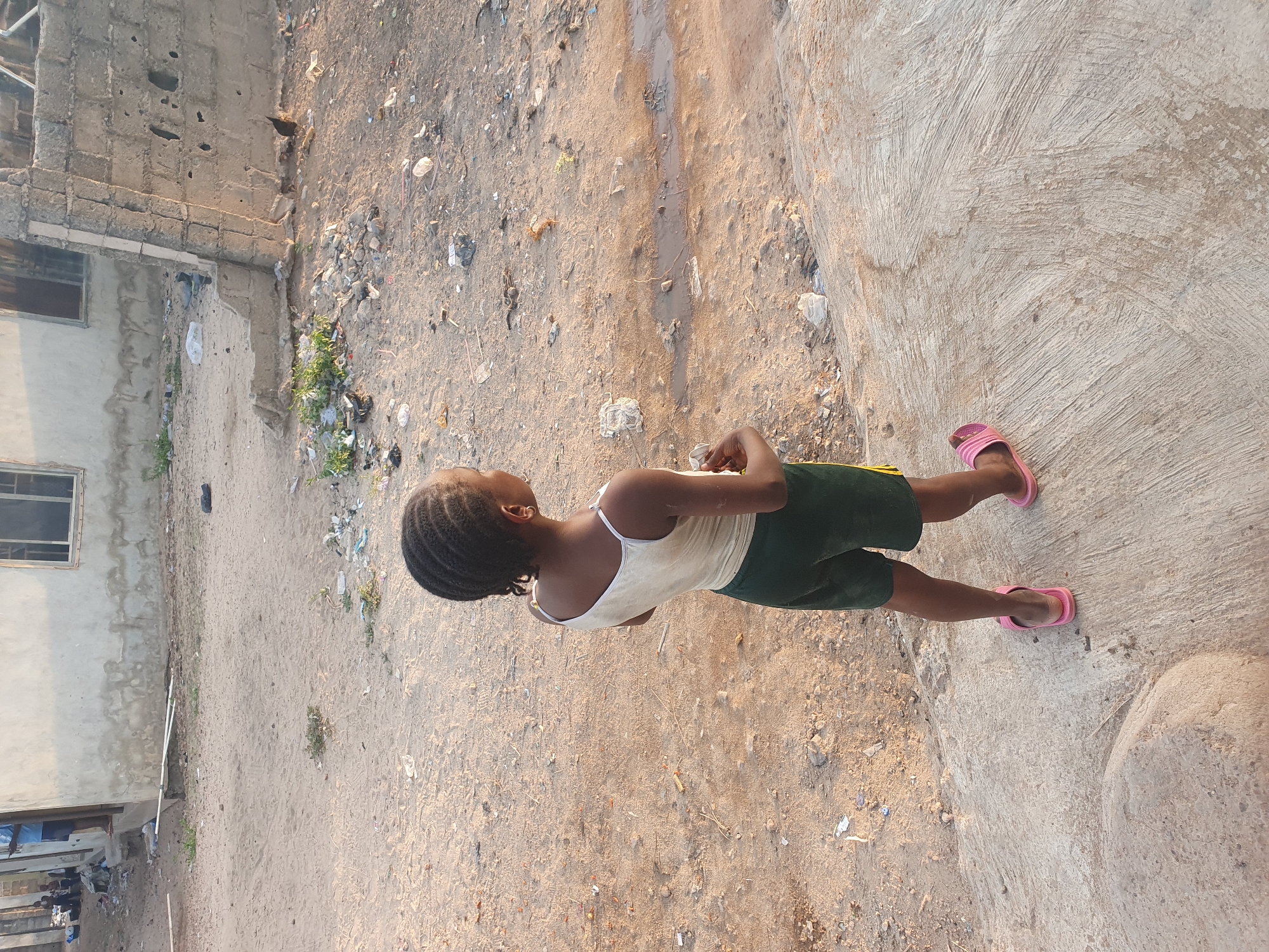 A young person, seen from behind and slightly to the right, stands on a mixed dirt and concrete surface under bright, warm sunlight. They have dark skin and braided hair, wearing a light-colored tank top, dark green shorts with a yellow stripe on the side, and pink flip-flops. Their arms are bent, with hands resting or clasped in front.
The ground is primarily light brown dirt or sand, heavily littered with scattered debris including plastic, paper, small stones, and twigs. A narrow, dark groove or track runs through the dirt. To the person's right, the surface changes to textured concrete with visible brush strokes.
On the left side of the frame, there are unpainted concrete block buildings, appearing somewhat unfinished or weathered. One building features a clear window with a dark frame. The scene depicts an outdoor, somewhat neglected urban or semi-urban environment, characteristic of parts of Lokoja, Nigeria, with visible signs of poor waste management. The strong, clear light suggests a sunny day, likely late morning or afternoon. No visible text is present.