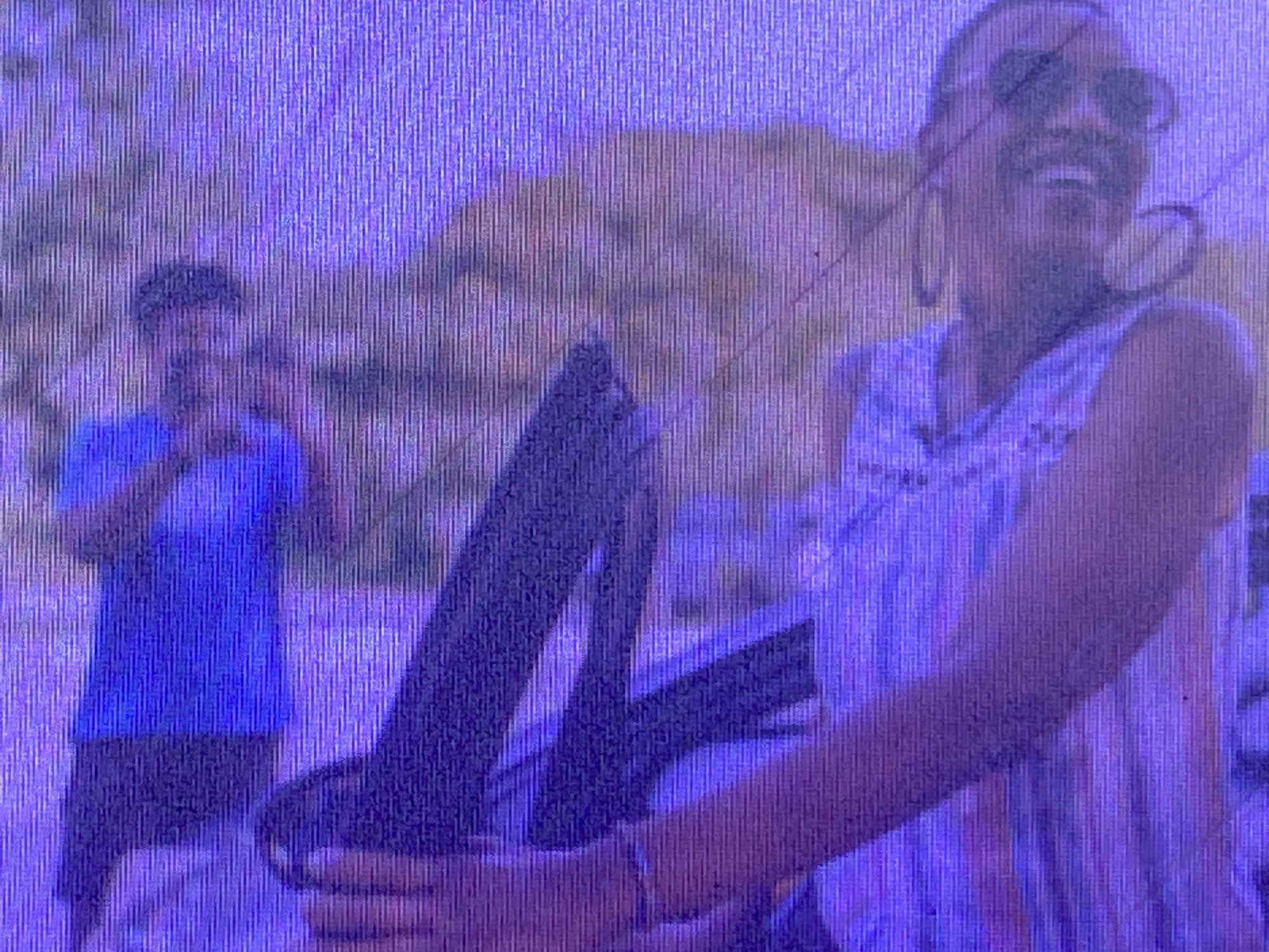 The media file depicts two individuals in an outdoor setting with a mountainous backdrop. The person in the foreground, closer to the camera, is wearing sunglasses and a sleeveless shirt. They appear to be smiling or laughing, with their mouth open. This individual is holding onto a dark-colored object, possibly part of a vehicle or some equipment. In the background, a second person is visible, wearing a blue t-shirt and holding something up to their face, possibly a camera or phone. The overall lighting suggests it might be daytime, and the presence of mountains hints at a location with natural topography. The grainy texture of the image suggests it may be an older photograph or a low-resolution digital capture. The context provided indicates the location is Ikorodu, Nigeria. No visible text is present in the image.