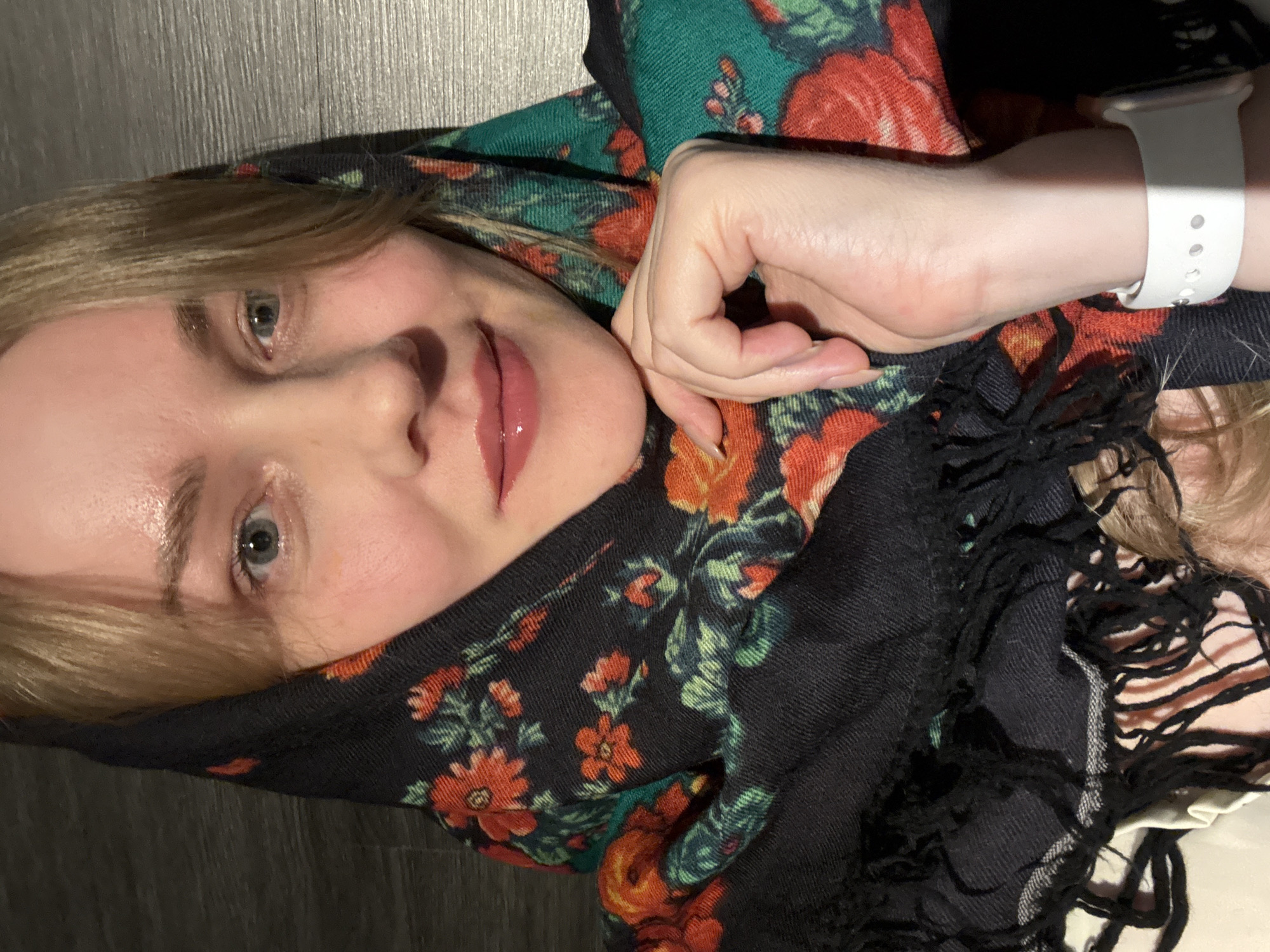 A person with blonde hair and fair skin is looking at the camera. They are wearing a colorful floral scarf, partially covering their face. The scarf is predominantly green and black with orange and red floral patterns. The person's hand is resting near their face, and they are wearing a white smartwatch. The background appears to be a dark wooden surface. The photograph's location in the city of Chaikovsky, Russia, is not possible to ascertain from this image alone. No actions or interactions are discernible.