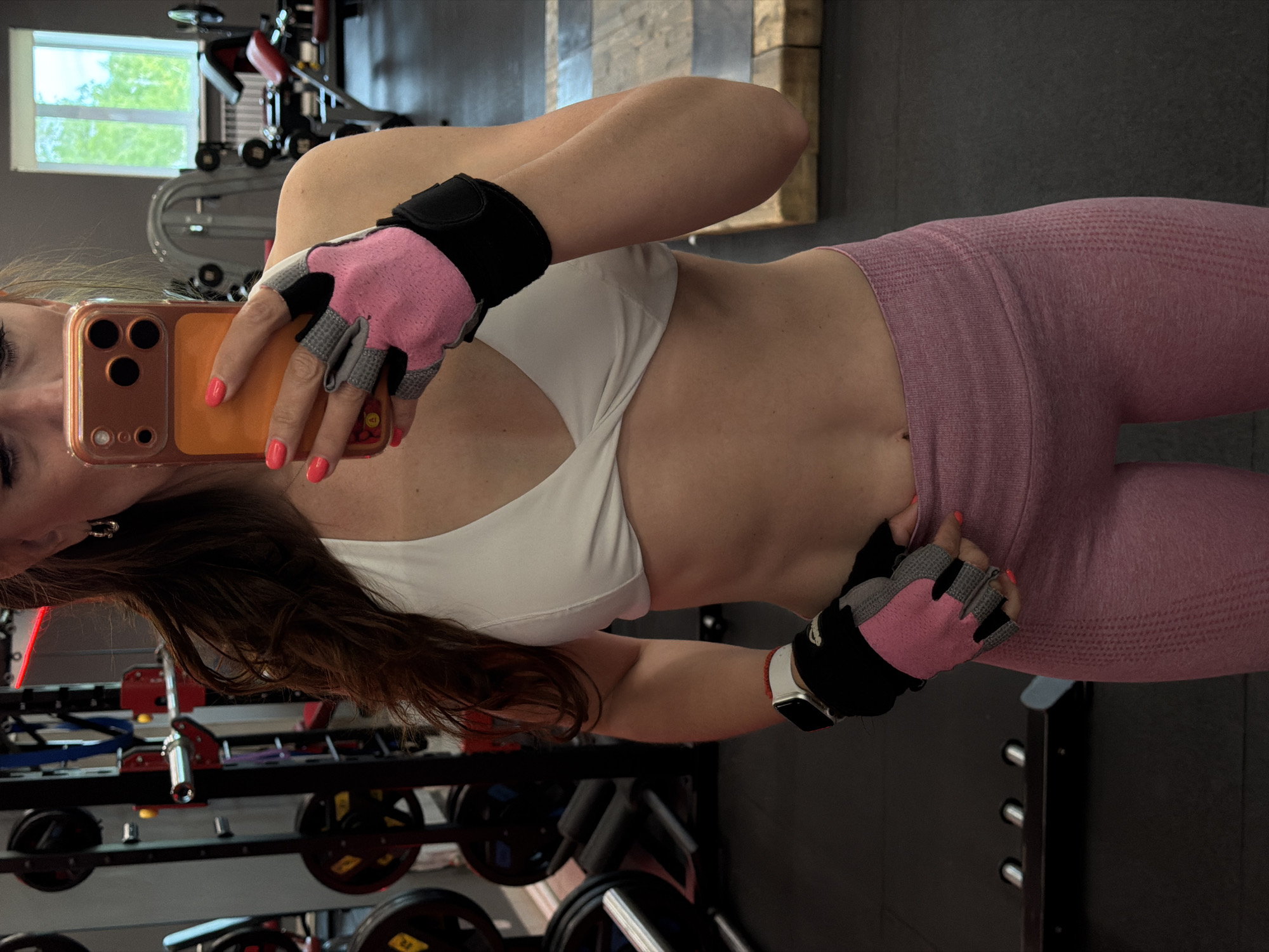 A woman is captured in a self-portrait within what appears to be a gym in Birobidzhan, Russia. The image is rotated 90 degrees clockwise. She is seen from approximately the waist up, holding a smartphone with an orange case in her right hand, positioned to take the photograph. Her fingernails are painted a bright coral, matching the phone case. She wears a white sports bra, form-fitting pink seamless-style leggings, and pink and grey fingerless weightlifting gloves on both hands. A white smartwatch is visible on her left wrist, which rests on her hip, gently pulling at the fabric of her leggings.
The background reveals a gym setting with various weight racks, barbells, and other exercise equipment. The floor is dark, and the walls are light grey. A rectangular window in the upper left corner shows green foliage outside, indicating it is daytime. The woman's long, dark brown hair flows down her back and shoulder. There is no clearly legible text on her clothing or the visible equipment.