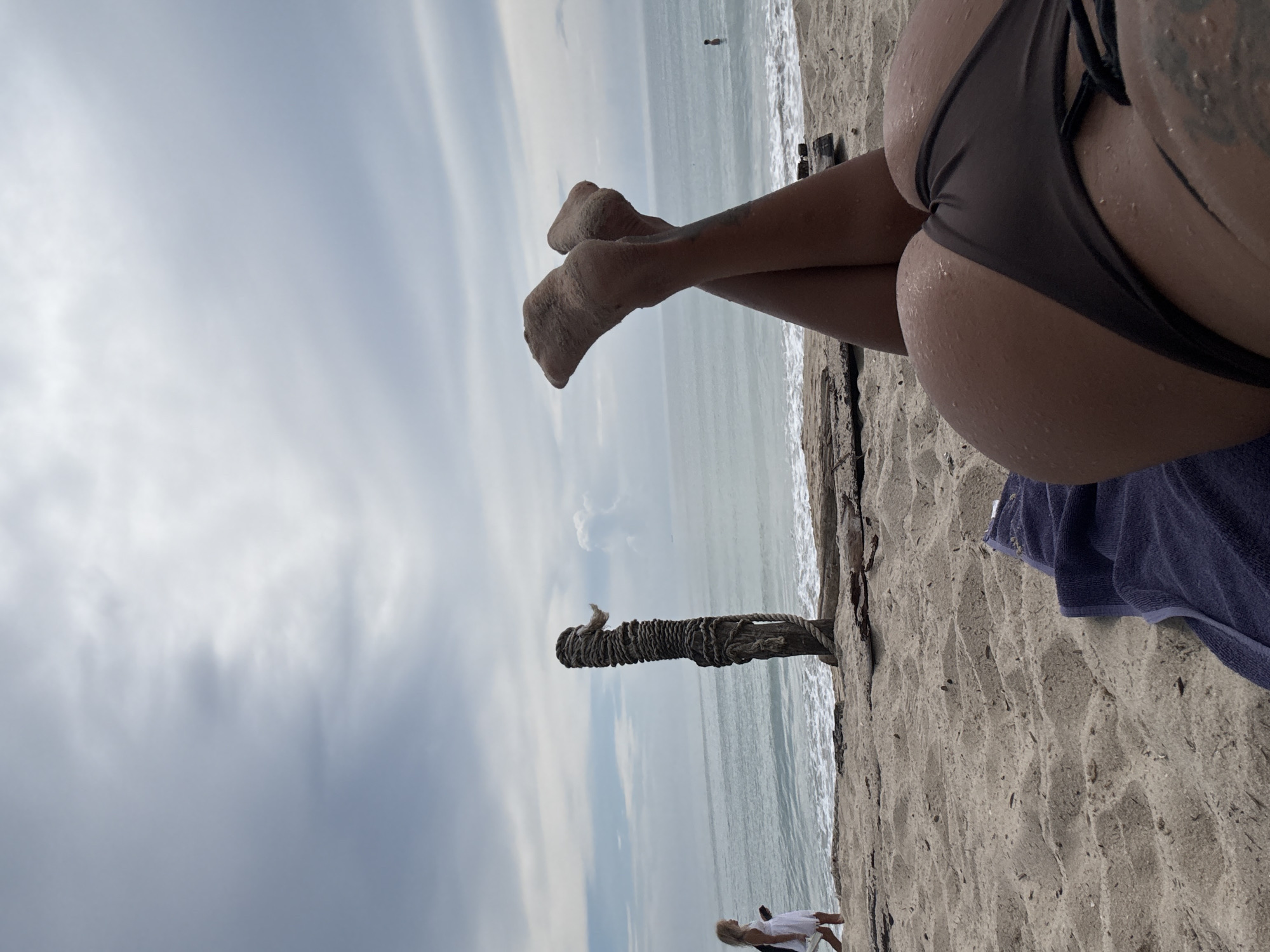 An extreme close-up shot shows the lower half of a person's body lying on a beach towel on the sand. Their legs are crossed at the ankles, and their feet are turned upwards. The skin is wet and sandy. In the background, the ocean meets a cloudy sky. To the left, a young girl in a white dress walks along the shoreline near a weathered wooden post wrapped in rope. A single person can be seen in the water in the far distance. The scene appears to be a beach in Choeng Thale, Thailand.