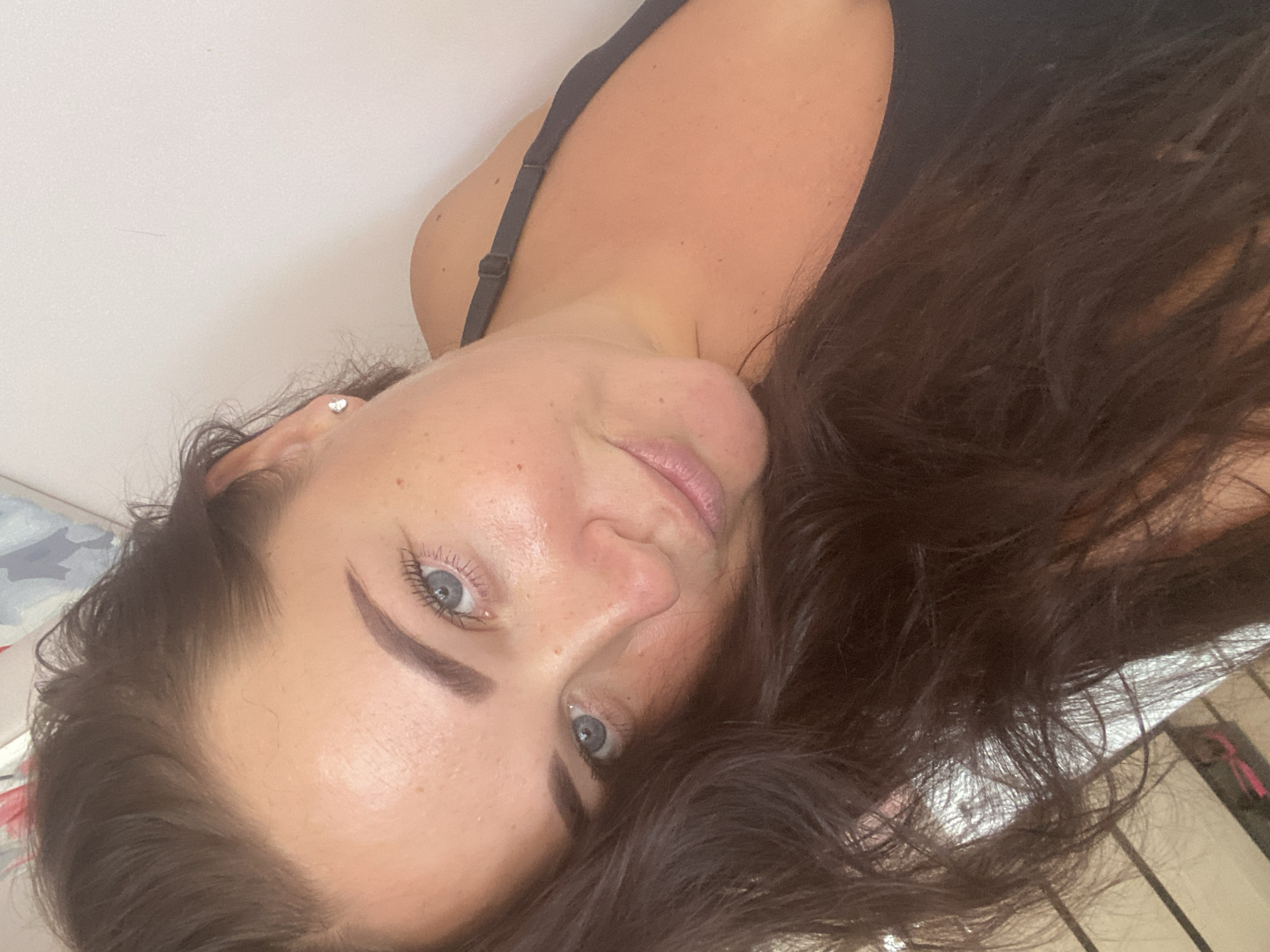 A woman with long, dark hair and blue eyes is taking a selfie. She is wearing a black tank top and a small stud earring in her left ear. Her skin is lightly tanned and has freckles. The background is a plain, light-colored wall and some blurry elements that suggest an indoor environment, possibly a bedroom or living area. The lighting is soft and natural. There is no discernible location in Sochi, Russia.