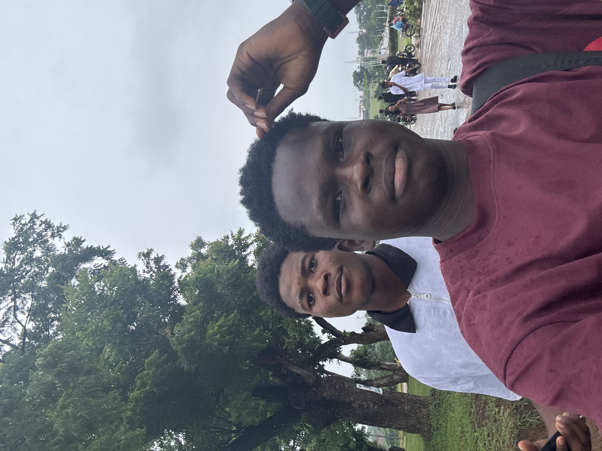 A portrait-oriented outdoor selfie captures two young dark-skinned men in the foreground, with an active urban scene and lush trees forming the background under an overcast sky.
The man positioned closest to the camera, on the right, has short, curly black hair and wears a maroon long-sleeved crew-neck t-shirt. A black backpack strap is visible over his right shoulder. His right hand, adorned with a dark green/teal watch featuring a square face, is raised to his head, touching his hair. A dark object, possibly a smartphone, is partially visible in his left hand at the bottom left of the frame. He looks directly at the camera with a neutral expression.
Behind and slightly to the left of the first man stands another young dark-skinned man, also with short, curly black hair. He is wearing a white short-sleeved collared shirt with a black collar and a white zipper pull at the front. His gaze is directed towards the camera with a neutral expression.
The left and central background is dominated by a dense cluster of large trees with verdant foliage and thick trunks, indicative of a heavily vegetated area or park. Grass is visible beneath the trees.
On the right side of the midground and background, a paved area, consistent with a road or thoroughfare, hosts several individuals and vehicles. A woman in a light brown dress is seen walking away from the camera, while a man in a white traditional Nigerian garment walks towards the left. Other indistinct figures are also present. Multiple motorcycles (okadas) are visible on the pavement, some parked, others with riders, suggesting active use. The pavement appears wet, possibly from recent rainfall.
The sky is uniformly overcast and light gray. In the far background on the right, distant low-rise buildings or structures are visible on a slight elevation, along with utility poles.
The overall environment, characterized by dense vegetation, the presence of motorcycles (okadas), and attire, is consistent with an urban or semi-urban setting in Nigeria. While the specific location within Ihiagwa, Nigeria, is not identifiable by explicit landmarks in the image, the scene aligns with the general characteristics of the region.