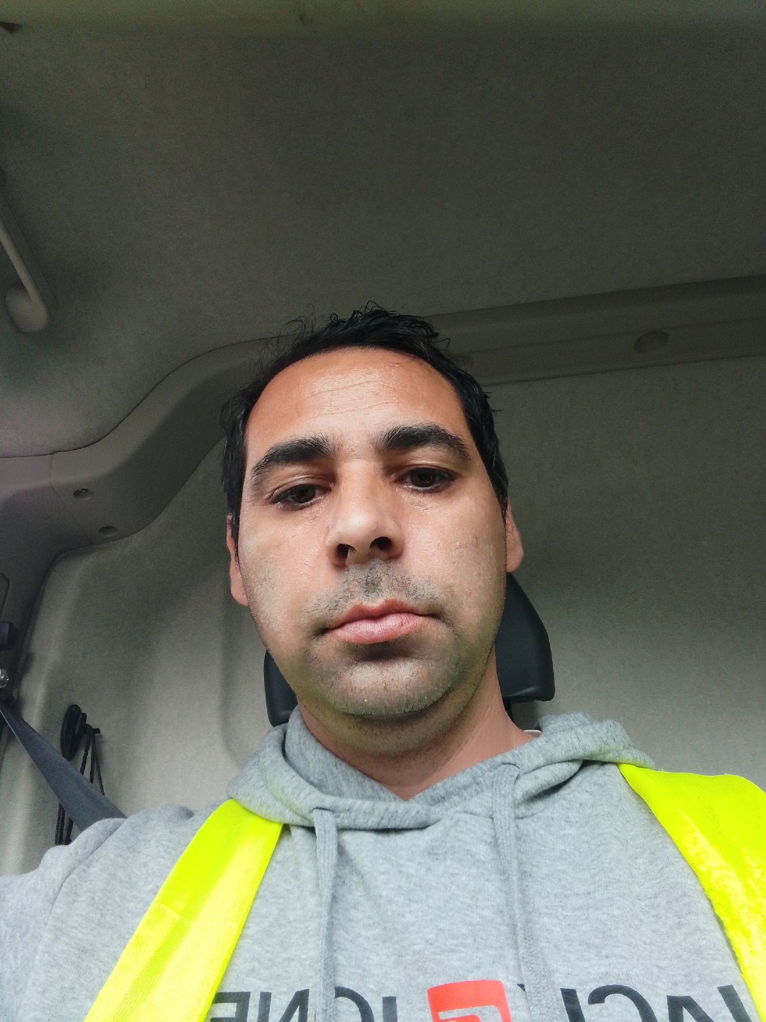 A man with dark hair and a light complexion is taking a selfie inside a vehicle. He is wearing a grey hooded sweatshirt with a bright yellow reflective vest over it. The interior of the vehicle appears to be a cab, with a dark headrest visible behind him and a seatbelt on his left. The background is a neutral, greyish interior of the vehicle. There are no discernible landmarks or indications of the city of Verden (Aller), Germany in the image.