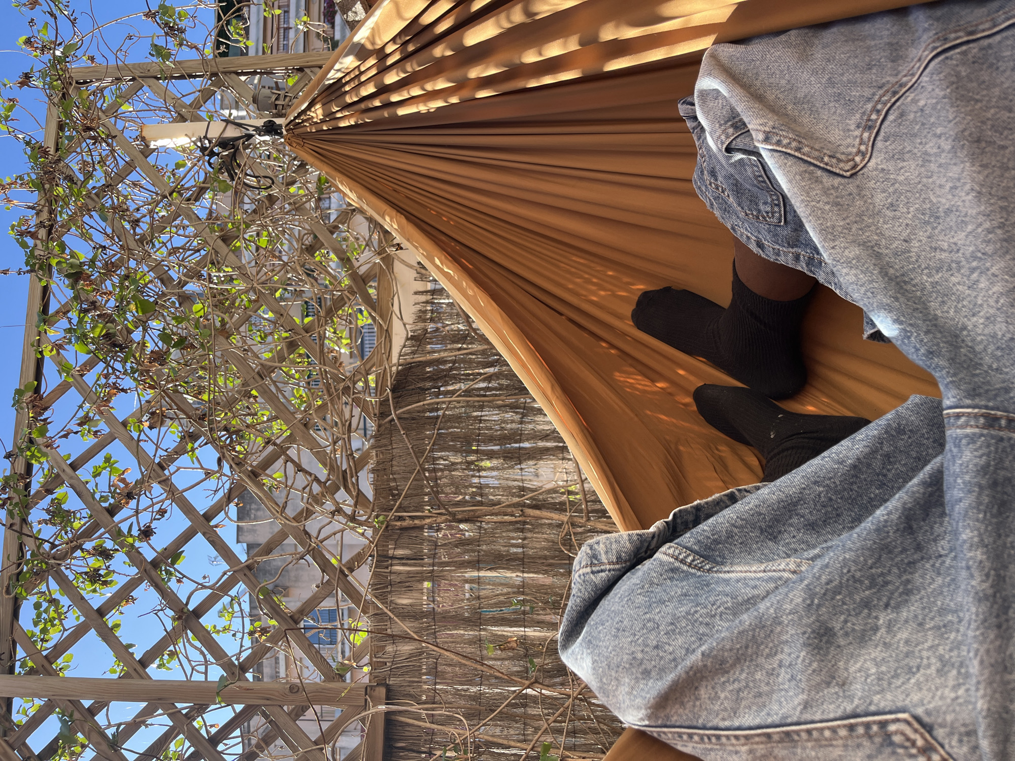 The image presents a view from within a brown hammock. A person wearing black socks and a light blue denim jacket is lying in the hammock. The hammock is strung up outdoors and is attached to a wooden lattice structure with climbing vines and foliage. In the background, buildings are visible through the lattice. The scene is bathed in natural daylight, with a clear blue sky visible. The photograph may have been taken in Barcelona, Spain due to the unique architecture in the background.