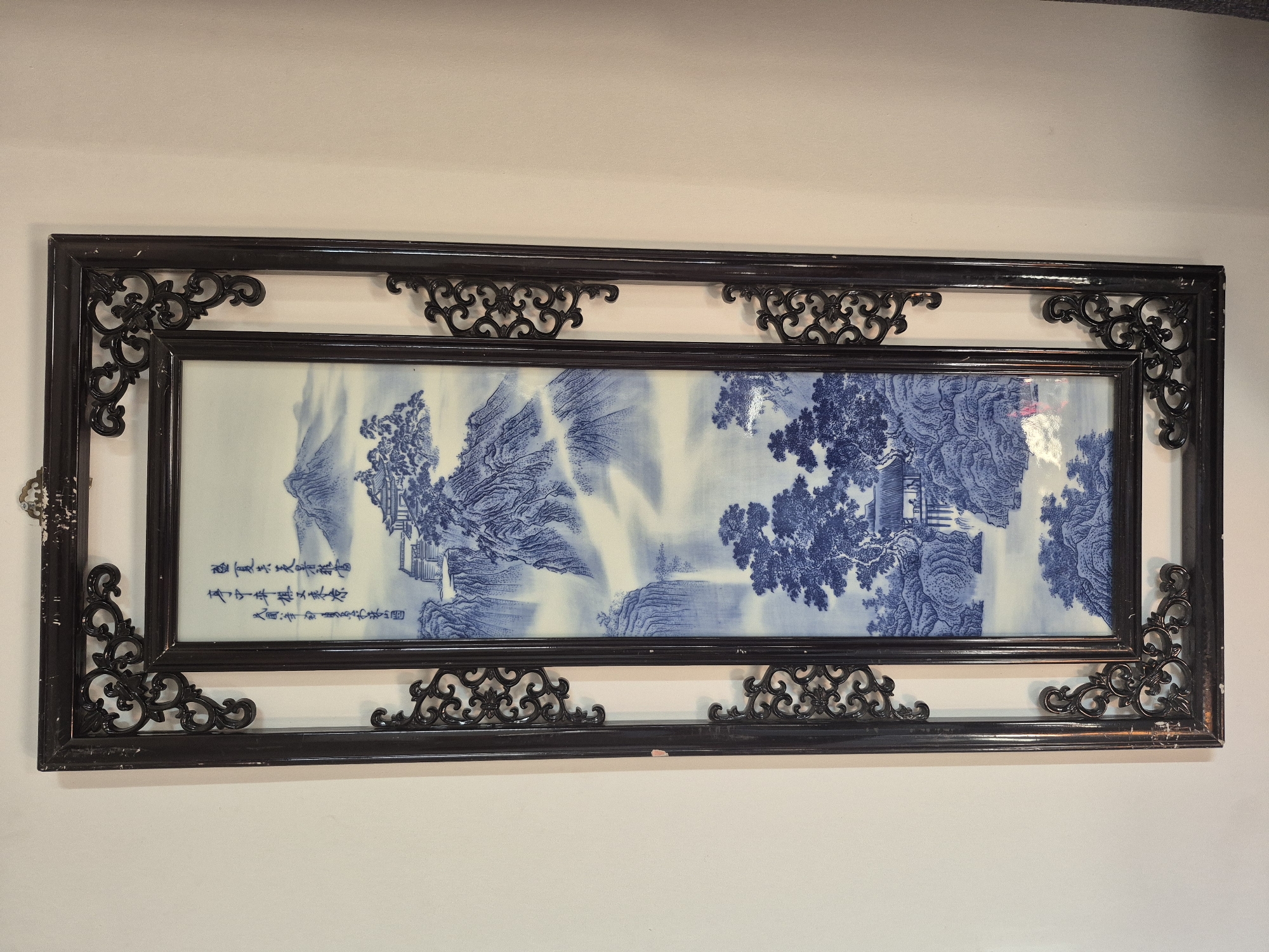 The image shows a framed piece of blue and white porcelain artwork depicting a traditional Chinese landscape with mountains, waterfalls, trees, and buildings. The frame is dark, intricately carved wood, with decorative scrollwork at the corners and along the sides. The artwork is hanging on a plain, light-colored wall, and the lighting suggests it is indoors. The image is a full shot of the artwork from a slightly elevated angle. There is no information in the image to suggest its location in the city of Prince Islands, Turkey.