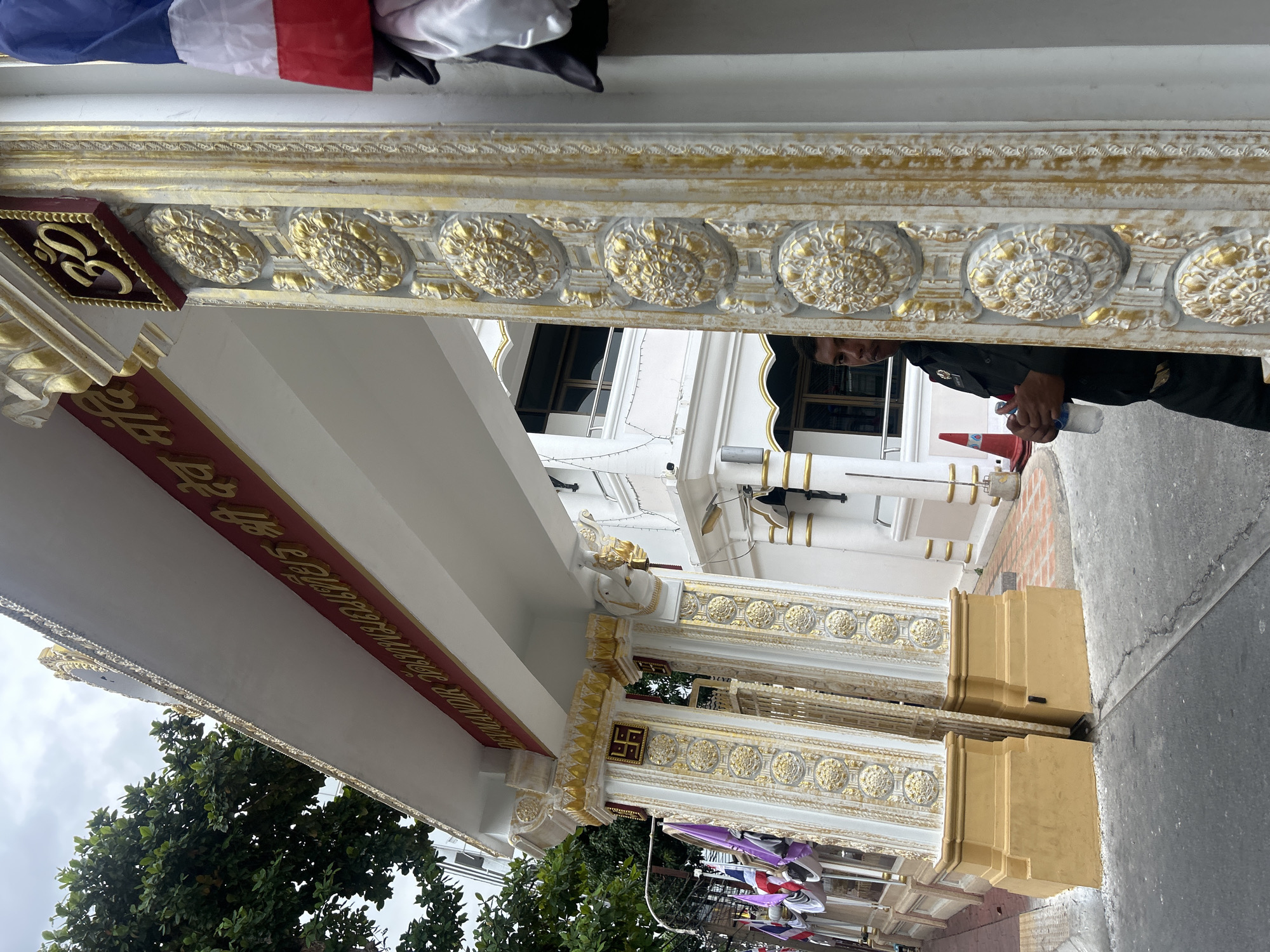 A man in a dark uniform holding a water bottle stands partially obscured by a decorative gold and white pillar. He is looking towards the left of the frame. Behind him is a white building with gold accents. Above the entrance, red signage with gold lettering is visible. Flags, including a Thai flag, are displayed on poles to the left of the pillar. The scene appears to be a street in Bangkok, Thailand, likely an entrance to a temple or religious institution. The sky is overcast.