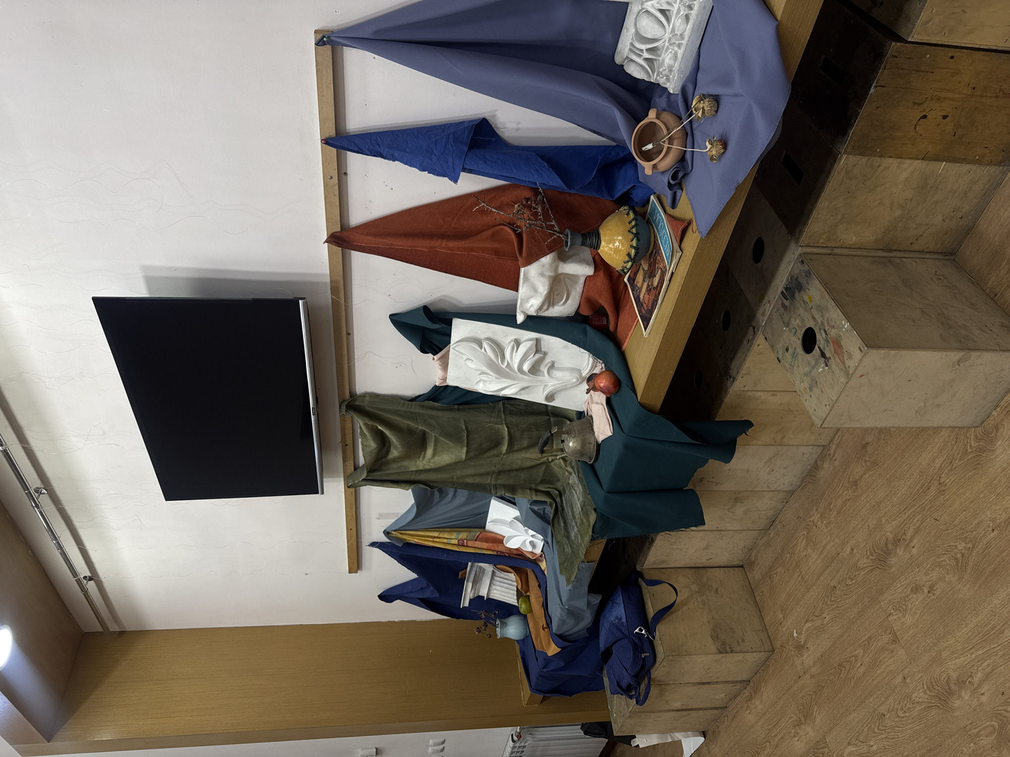 The image shows a still life setup indoors against a white wall with a television mounted on it. The still life is arranged on a wooden shelf or ledge. Various draped fabrics in colors like blue, brown, green, and gray serve as backgrounds.
Objects included in the still life are a white plaster ornament, a small clay pot, a yellow and blue vase, a book, a piece of fruit (likely an apple), a metal vessel, and other assorted items partially concealed by the fabrics.
The scene is lit by overhead artificial light. Below the still life, a staircase-like structure is made of wooden boxes. On the floor near the base of the structure, a blue backpack is visible.
The image does not contain any indications about the location in Yerevan, Armenia.