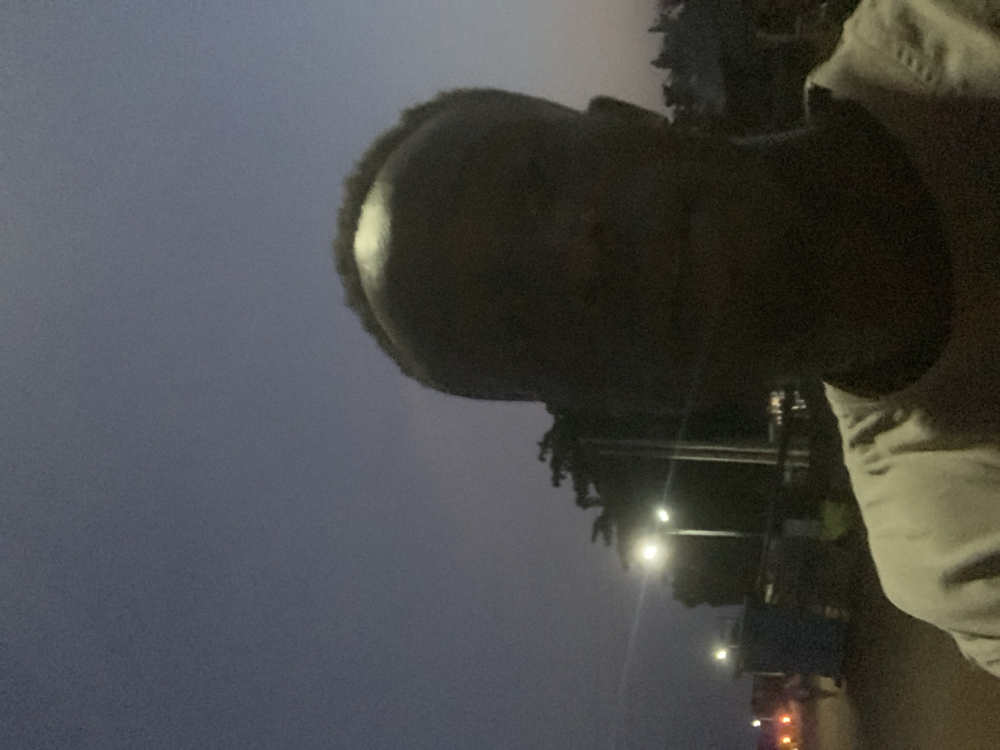 A selfie photograph taken at dusk or night in Gbeleko, Nigeria. The person in the image has dark skin and is wearing a light-colored shirt. Streetlights are visible in the background. The sky is dark.