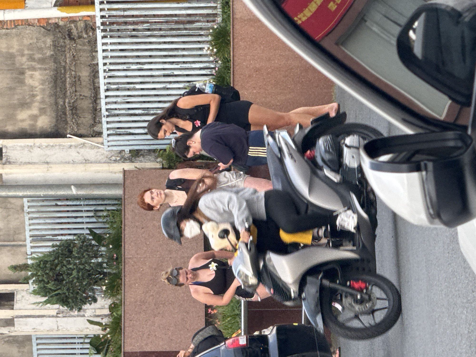 Four young women are pictured outdoors in what appears to be a city setting in Da Lat, Vietnam. The foreground is dominated by a silver scooter with a person wearing a helmet and a face mask, and possibly a child behind them. To the left of the scooter, a black car is visible, with the edge of its rear bumper and tail light showing.
Behind the scooter, three young women stand together. The woman on the far left wears a black tank top and shorts, with her hair pulled up and sunglasses on her head. She is looking down. The woman in the center is wearing a gray long-sleeved shirt and black pants, and is riding the scooter. The woman on the far right is wearing a black tank top and shorts.
Further in the background, there is a textured wall and a metal fence. To the left, a small tree is planted in a planter box. The overall impression is a snapshot of everyday life in a Vietnamese city.