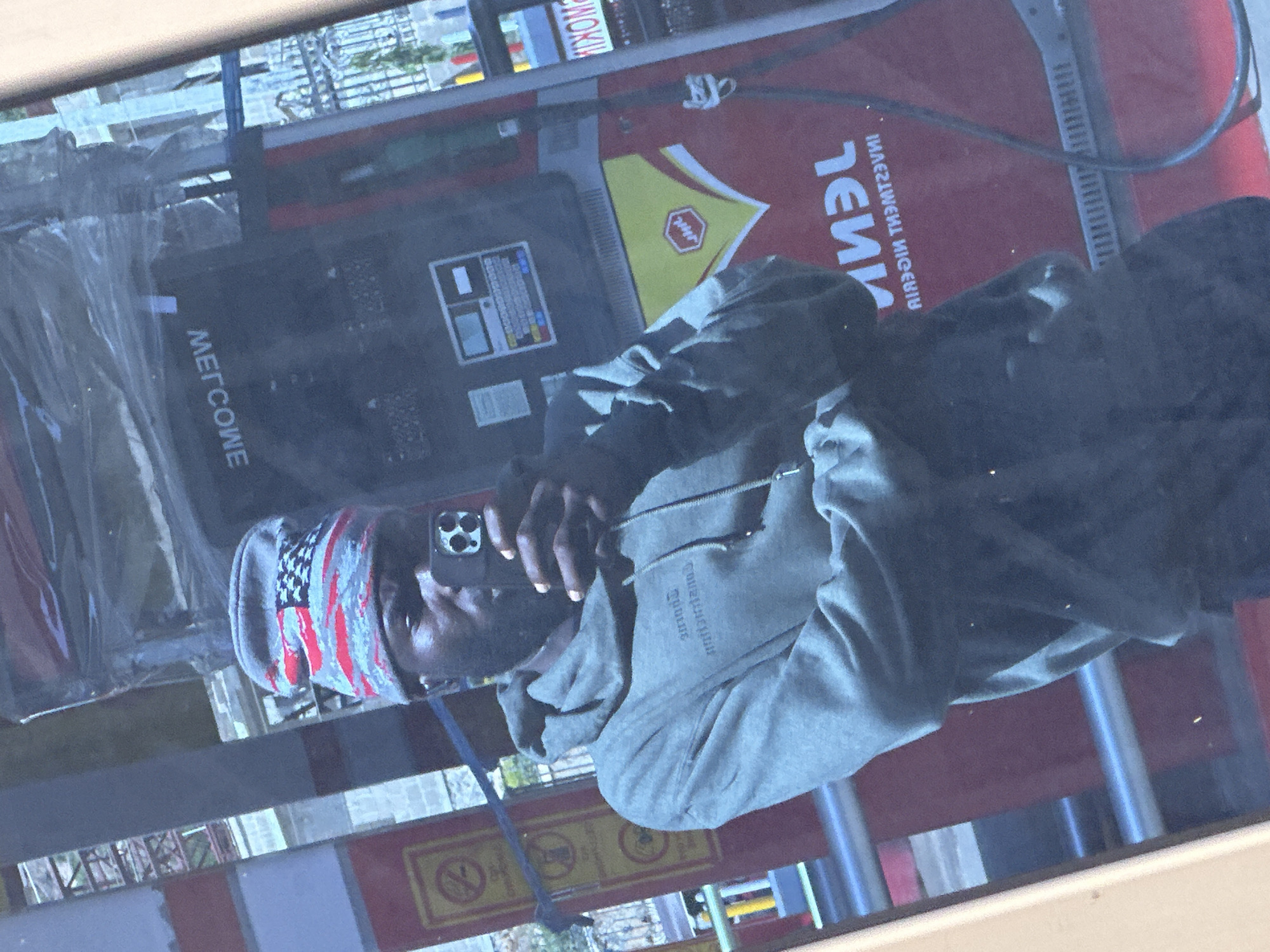 A person is taking a selfie in front of a gas pump. The person is wearing a beanie with an American flag design and a grey hoodie. The gas pump has a screen with text that reads "WELCOME" and a logo with a red and yellow design. In the background, there are reflections of buildings and trees, suggesting an outdoor urban environment. The gas pump is likely located at a gas station in Gura Top, Nigeria, as indicated by the surrounding context.