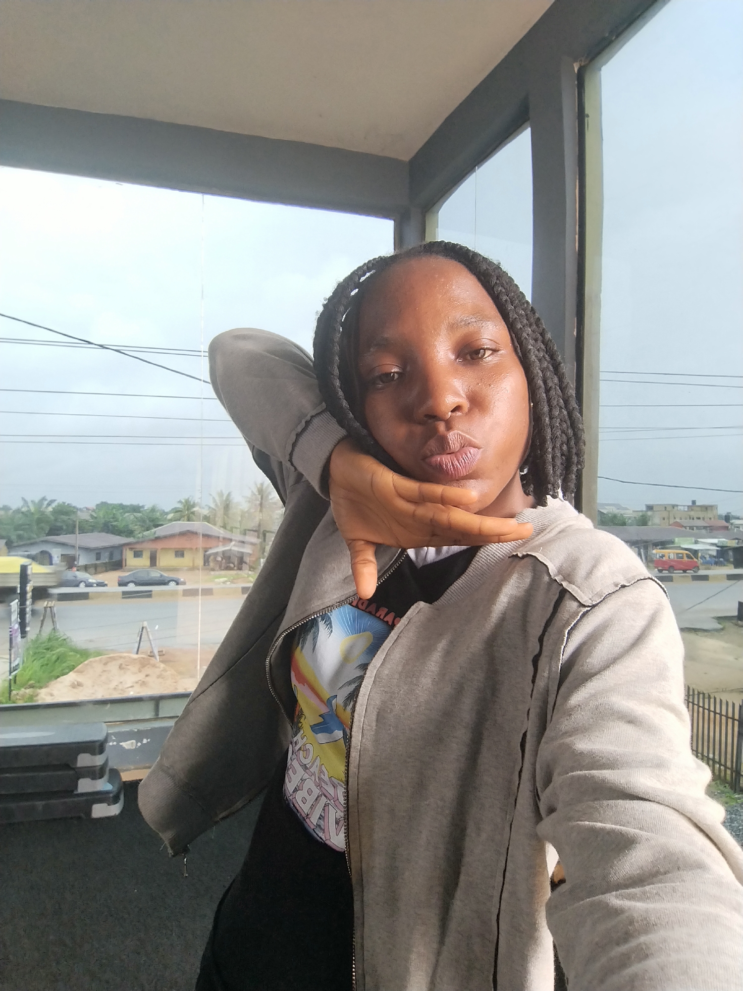 A young Black woman with braided hair is posing for a selfie indoors, with her left arm bent at the elbow and her hand resting on her cheek, pursing her lips. She is wearing a grey hooded jacket over a black t-shirt with a colorful graphic print. The background shows a large window with a view of a street scene in Uselu, Nigeria. Buildings, palm trees, and vehicles are visible outside. Electrical wires are strung across the sky. Inside, to the left of the woman, a stack of grey aerobic step platforms is visible. The lighting suggests it is daytime, possibly overcast.