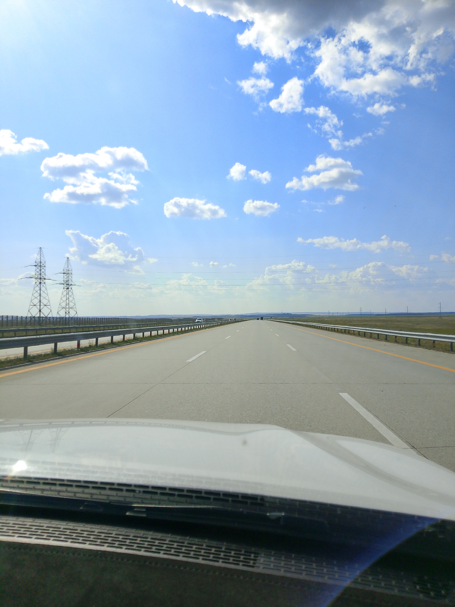 The image depicts a highway scene in Makinsk, Kazakhstan, under a bright, cloudy sky. The perspective is from inside a vehicle, looking forward through the windshield. A grey car hood is visible in the foreground, reflecting the sky and surrounding landscape. The highway itself is paved with asphalt and marked with white lines. On either side of the highway are metal guardrails and grassy fields. In the distance, two large metal power line towers stand prominently against the horizon, with additional power lines stretching across the landscape. A few cars are visible on the highway in the distance. The overall environment suggests a vast, open, and somewhat desolate landscape, typical of the steppes in Kazakhstan.