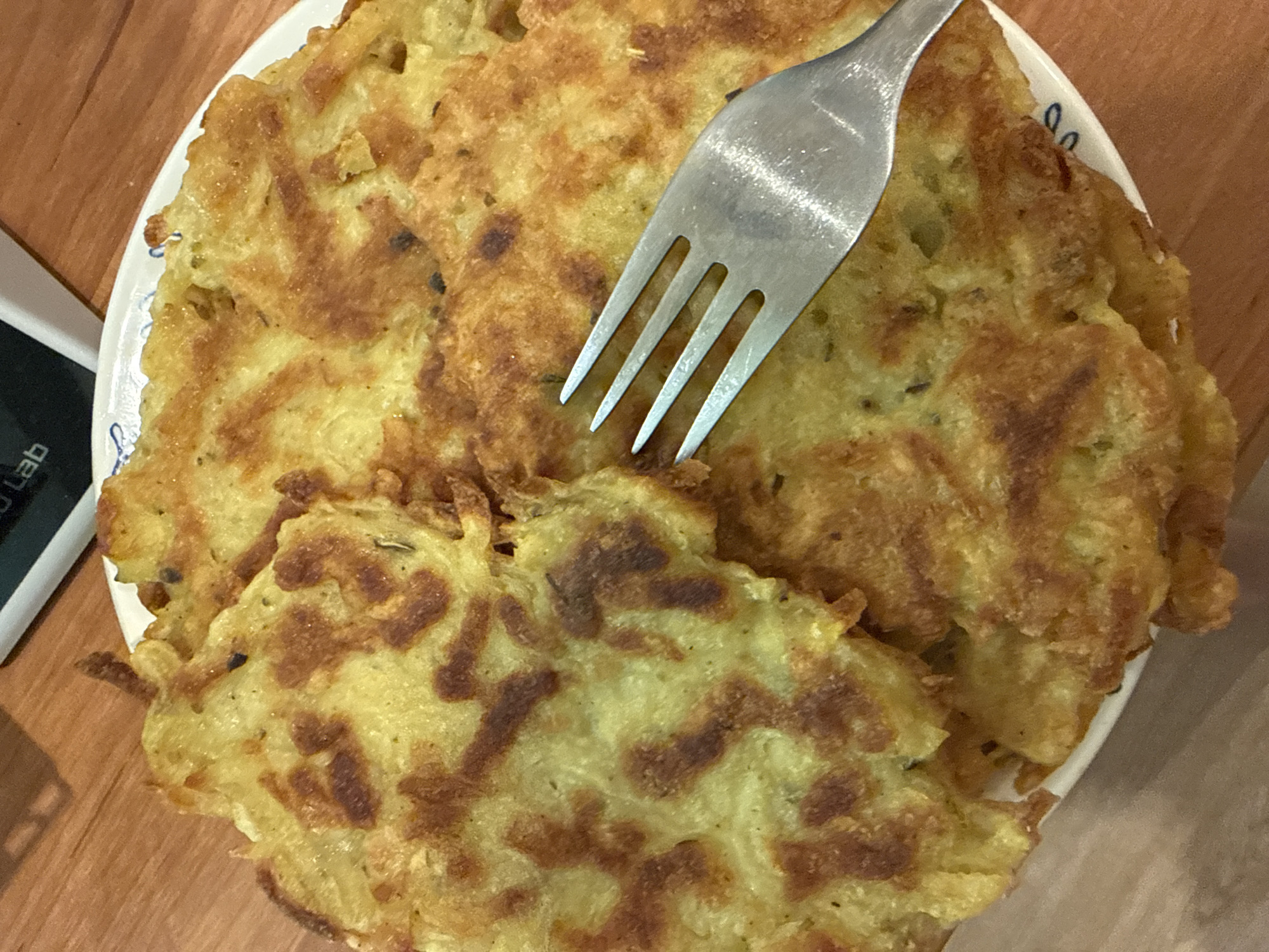 A top-down, close-up shot shows a white plate filled with potato pancakes, one of which is being pierced by a silver fork. The plate, decorated with small blue designs around the rim, sits on a wooden surface. A portion of a black electronic device, partially obscured, lies next to the plate on the left, with the word "Lab" visible. The pancakes are golden brown, with darker brown patches scattered across their surfaces, indicating cooked edges. The fork is positioned in the center of the frame, with its tines penetrating a large pancake.