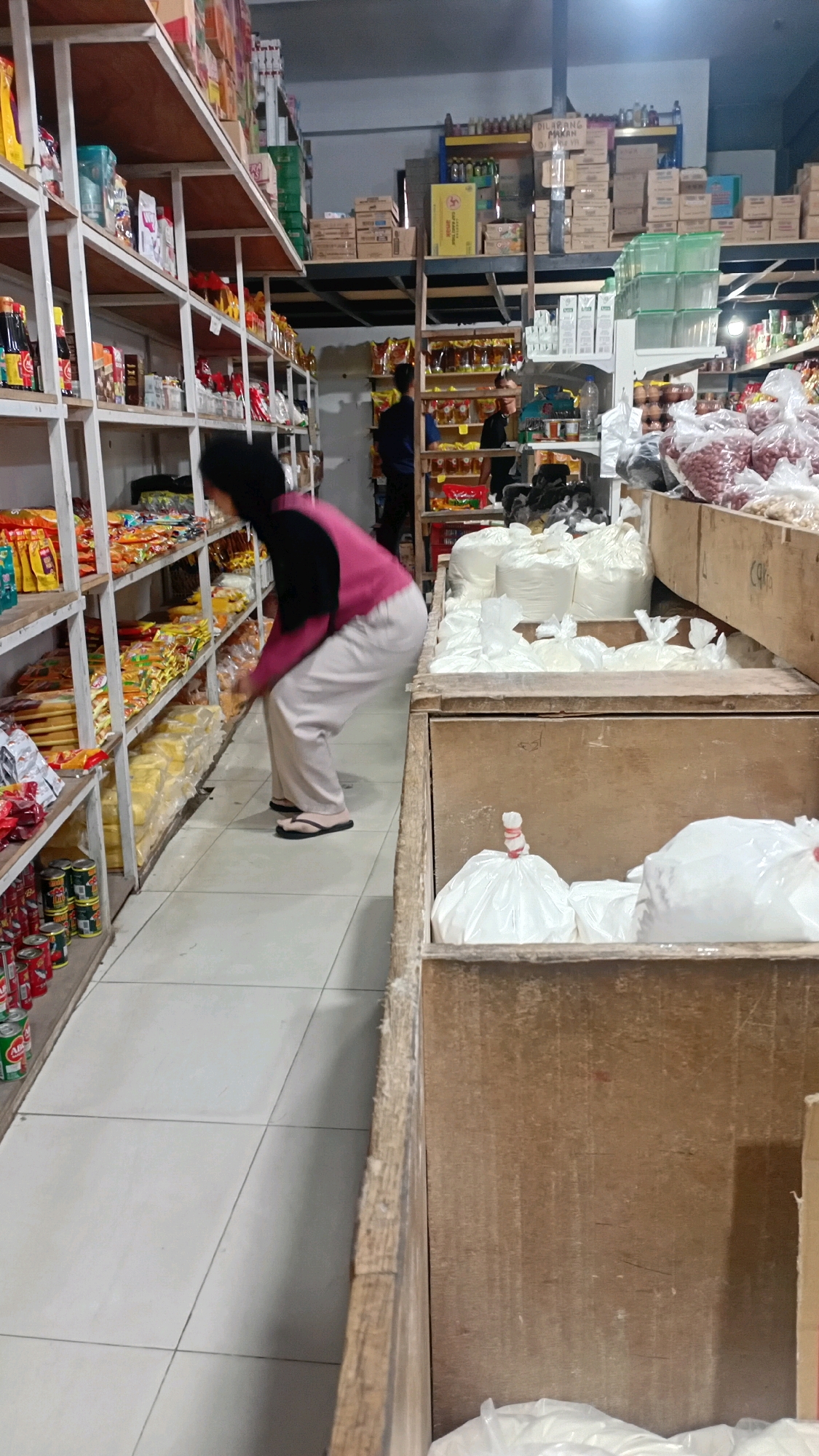 A woman wearing a black hijab and a pink long-sleeved shirt with light-colored pants is bending down in an aisle of a grocery store. The aisle is lined with shelves filled with various packaged goods, including cans and bagged items. To the right of the aisle are large wooden crates filled with white bags, likely containing bulk food items. Another person is visible in the background, partially obscured by shelves and products. The store appears to be well-stocked, with a variety of products displayed on multiple levels of shelving. The floor is tiled.