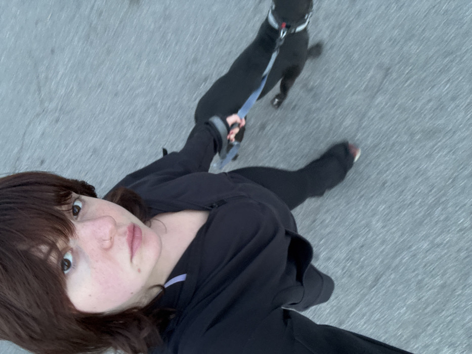 A selfie captures a person with brown hair and bangs, gazing towards the camera. They wear a black jacket and pants, likely on a walk. A black dog, leashed and collared, is positioned further ahead and to the right. The background consists of gray pavement, indicating an outdoor setting. The angle suggests the photo was taken from a slightly elevated perspective, looking downward. The individual appears to be in motion.