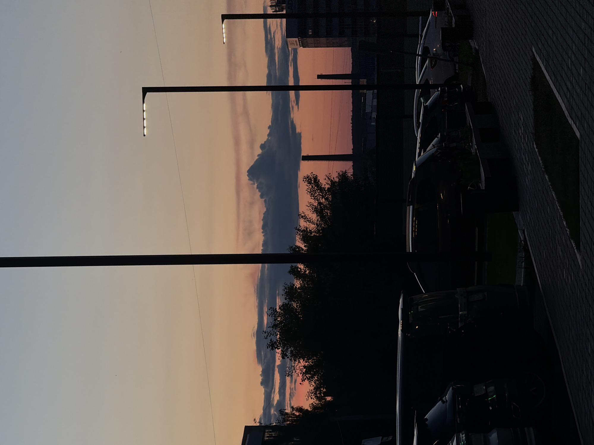 Captured at dusk, the image presents a street scene likely situated in Izhevsk, Russia, based on discernible architectural cues. Several dark-colored vehicles are parked alongside a sidewalk composed of grey patterned brick. Three modern streetlights with LED arrays illuminate the area. The background shows buildings including a high-rise with illuminated windows, the sky exhibits a gradation of colors, ranging from light orange to a muted violet, with clouds streaking horizontally across the horizon. Silhouetted trees partially obscure the view between the streetlights and buildings.