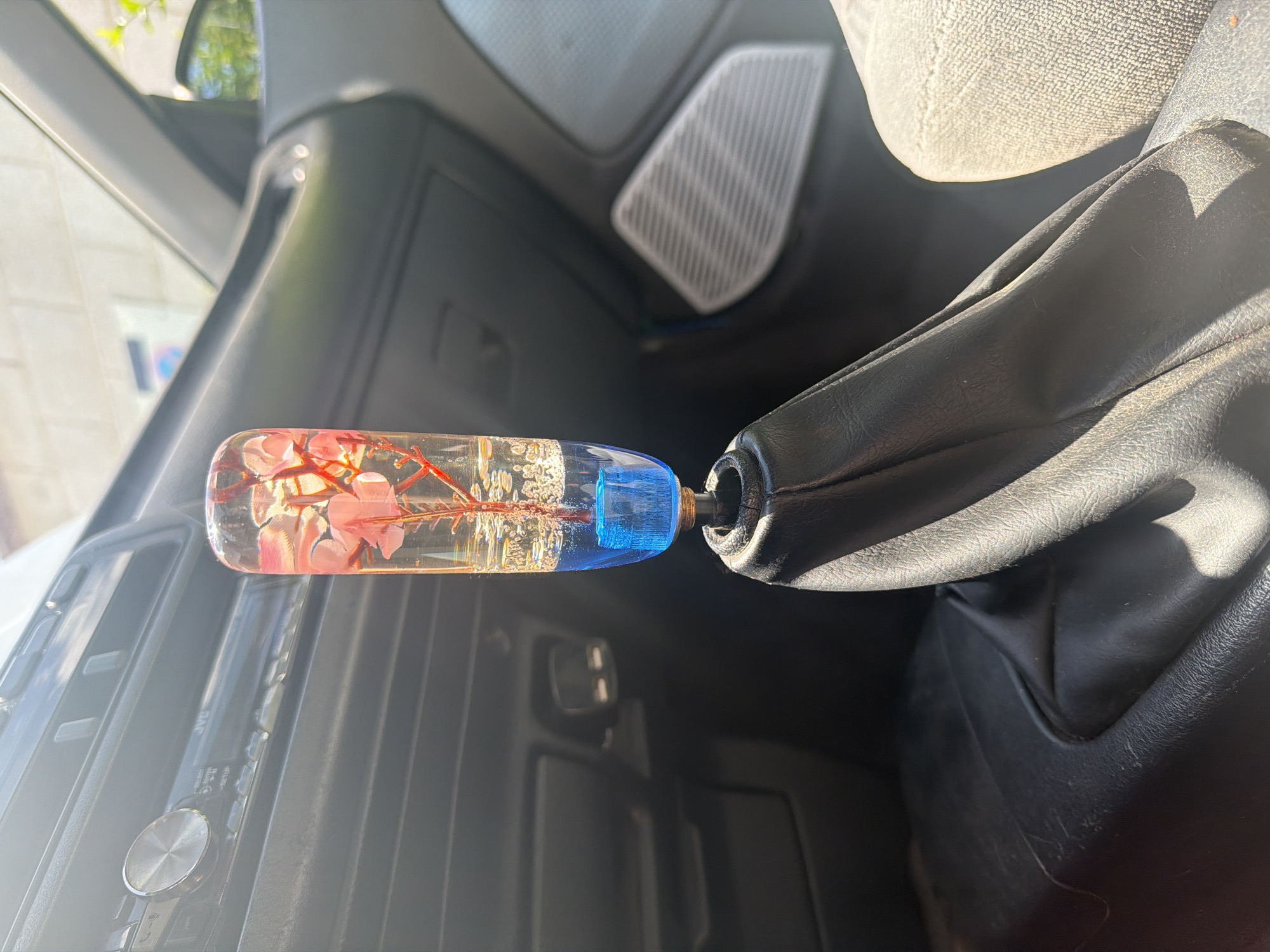 The image shows the interior of a car. The focus is on the gear shift, which has a custom shift knob made of clear resin, encapsulating pink flowers and possibly other botanical elements. The base of the knob is blue. The gear shift boot is made of black leather. The car's dashboard is visible in the background, along with a JVC car stereo. The interior appears to be older, as the dashboard style is from an older car. The environment suggests the image was taken outdoors during daylight, given the sunlight shining into the vehicle. The specific location in Varna, Bulgaria cannot be determined from the image.