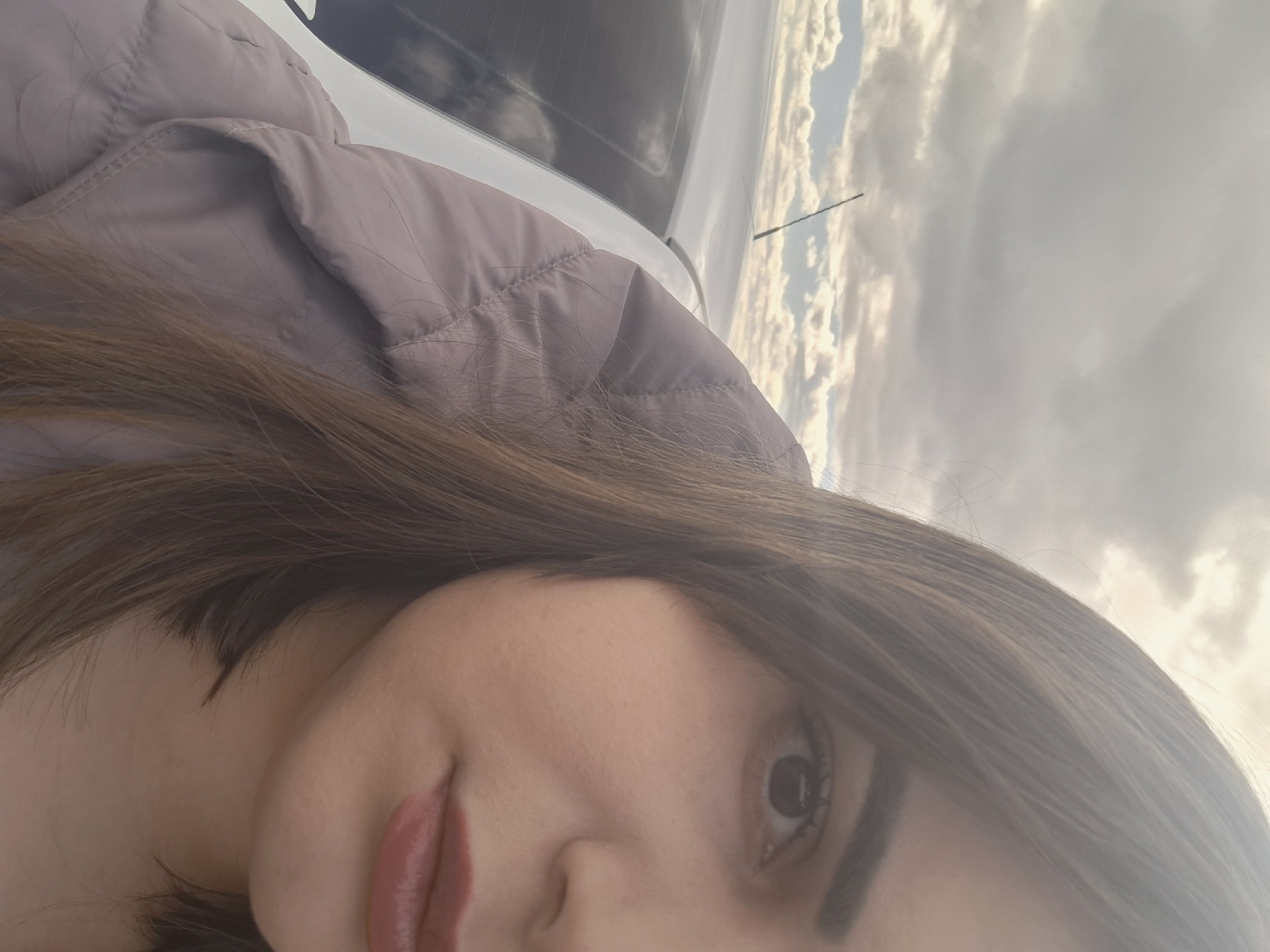 A close-up selfie of a woman with brown hair, light skin, and pink lipstick. She is wearing a light purple jacket. The background shows a car and a cloudy sky. The location is not possible to determine only by this picture, the city of Povarenki in Russia cannot be verified.