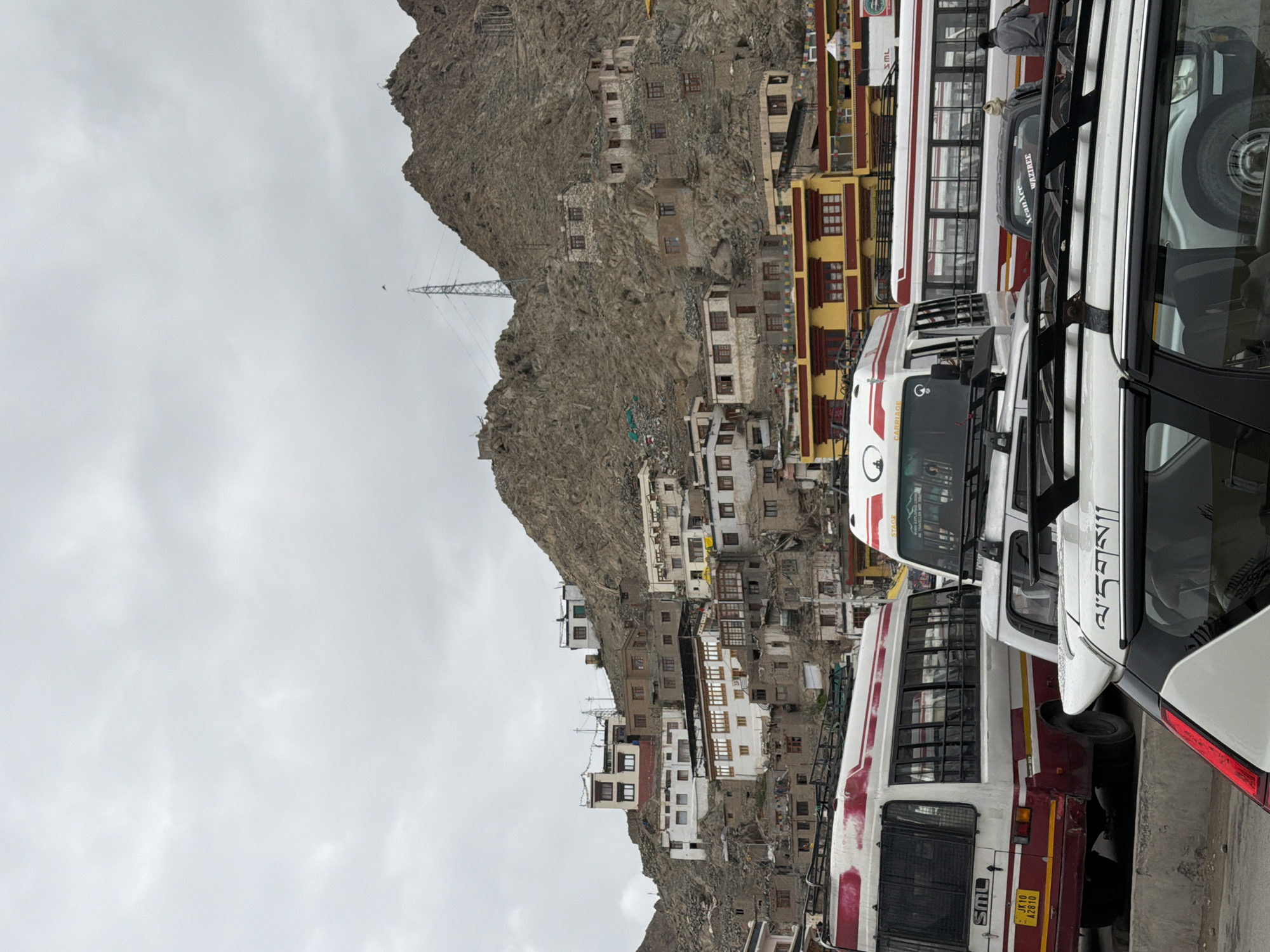In the city of Leh, India, a cloudy day is captured with a view of buildings constructed on a rocky mountainside. The buildings are predominantly made of stone and plaster, with some featuring white facades and brown-tiled roofs. Several buses are parked in the foreground, including a white and maroon bus with the license plate "JK10 A2810." Behind the buses, a cluster of yellow and red buildings is visible, adorned with prayer flags. A person in a grey jacket is walking away from the camera in the middle ground. The overall scene depicts a densely populated mountainside settlement with various modes of transportation.