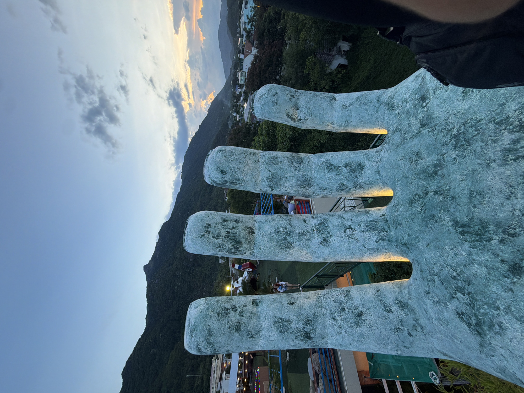 This is a close-up, upward-angle, medium shot of a large, textured, turquoise-colored statue resembling fingers of a hand. The statue is positioned in the foreground, dominating the lower half of the frame. The surface of the statue appears rough and somewhat mottled. Behind the statue, in the mid-ground, there is a multi-level outdoor recreational area with railings and some people visible on walkways and platforms. In the background, a densely vegetated hillside rises, with scattered buildings and residential areas dotting its lower slopes and extending towards a distant mountain range. The sky above is a gradient of dusk colors, with a clear expanse of pale blue at the top transitioning to hues of orange and yellow near the horizon, punctuated by scattered, greyish clouds. The scene is likely from the Ba Na Hills Golden Bridge near Da Nang, Vietnam, given the iconic hand statues and mountainous landscape.