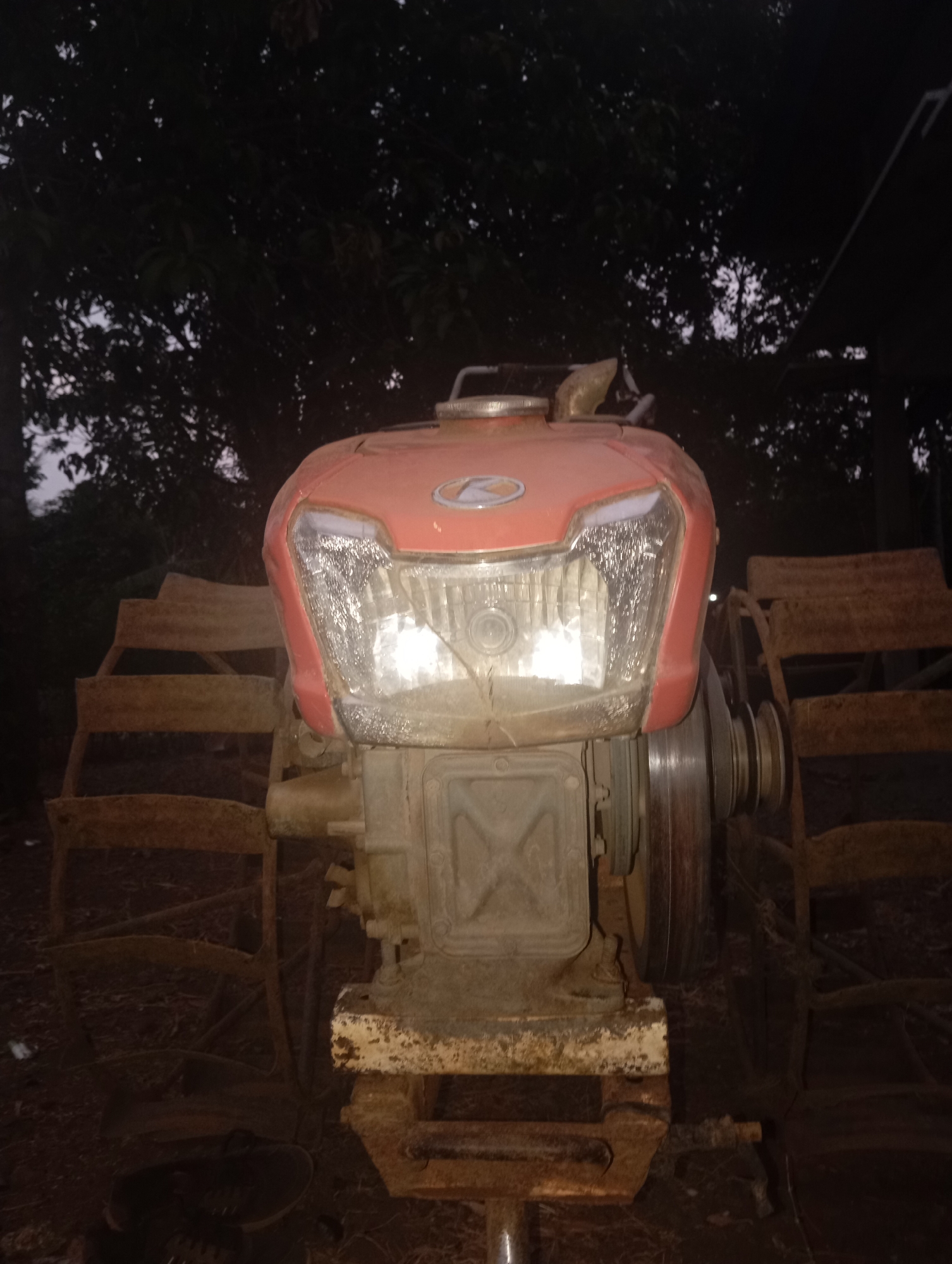 An aged, red-colored, two-wheeled tractor is positioned centrally in a dimly lit outdoor environment. The tractor features a prominent, modern-looking headlight assembly. Flanking the tractor are two large, spoked metal wheels that appear to be designed for rough terrain. The surrounding area is indistinct, with dark foliage visible in the background, suggesting a rural or semi-rural setting at dusk or night. A single black shoe lies on the ground in the lower-left quadrant of the image. The overall scene lacks activity and appears static. The location is identified as Kedungbenda, Indonesia.