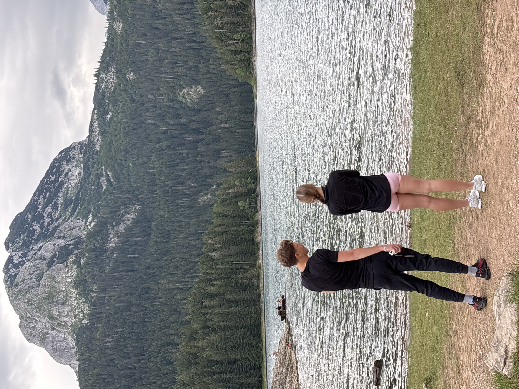 In Pitomine, Montenegro, two individuals, a male and a female, stand on a grassy shore facing a large body of water. The male, on the left, is wearing a black t-shirt and black pants, and is looking to his left. The female, on the right, is wearing a black long-sleeved shirt and pink shorts, and is looking out at the water. In the middle ground, several people are in a small wooden boat on the lake. The background features a dense forest of evergreen trees leading up to a large, rocky mountain under a cloudy sky.