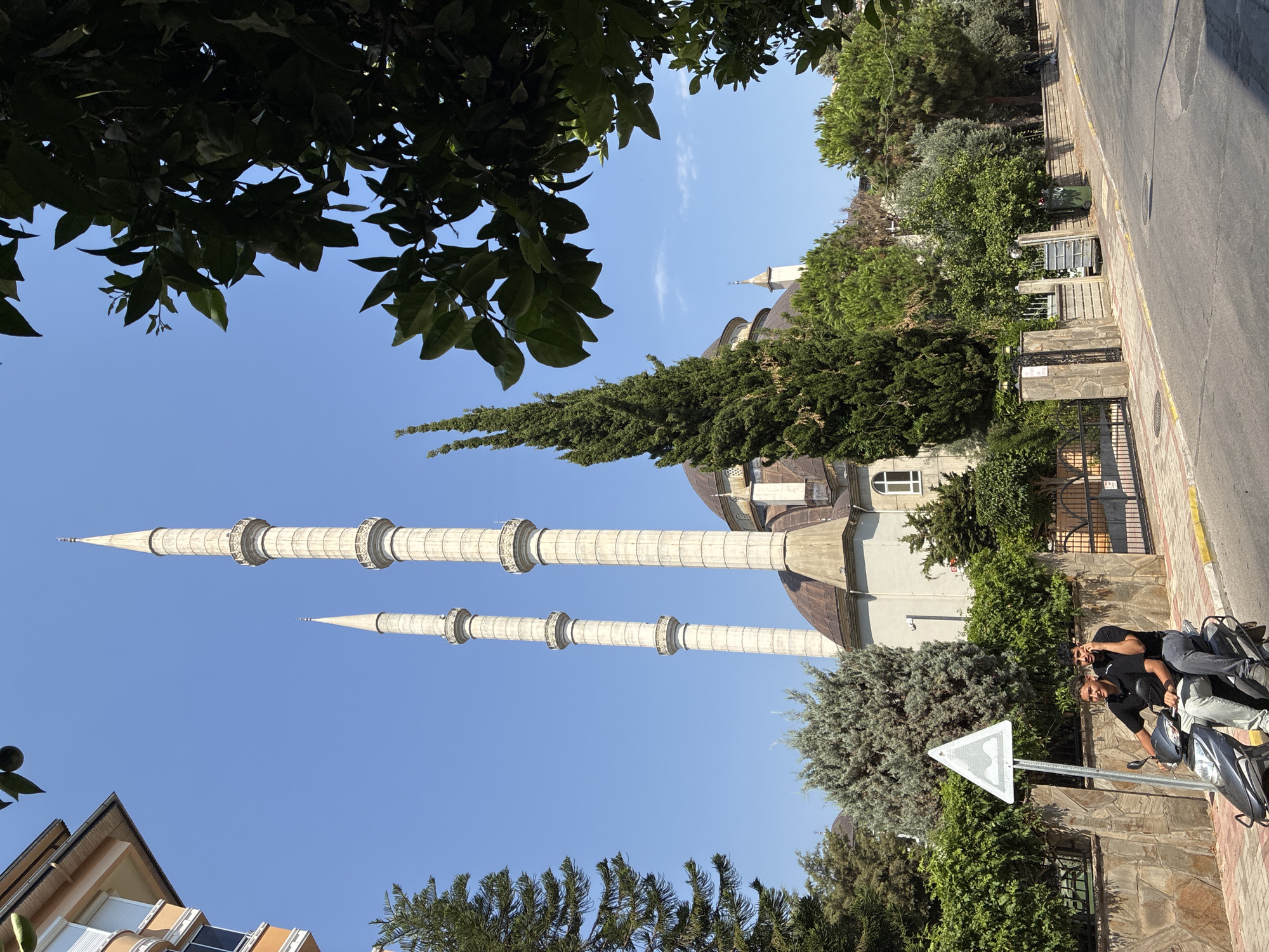 The image captures a street scene in Alanya, Turkey, dominated by a mosque with two tall, slender minarets. The mosque's main structure features a dome and is flanked by lush green trees and foliage, characteristic of the region. In the foreground, two young men are riding a scooter along a paved road. The man in the front is wearing a black polo shirt and light-colored pants, while the passenger behind him is dressed in a black shirt and dark jeans. They are both looking forward, seemingly engaged in conversation or focused on their ride. A triangular road sign with a graphic of two humps is visible near the scooter, indicating a speed bump ahead. The environment is bright and sunny, with a clear blue sky. The buildings in the background, including an apartment building to the left, suggest an urban setting. The overall impression is a snapshot of daily life in Alanya, with a blend of architectural heritage and modern transportation.