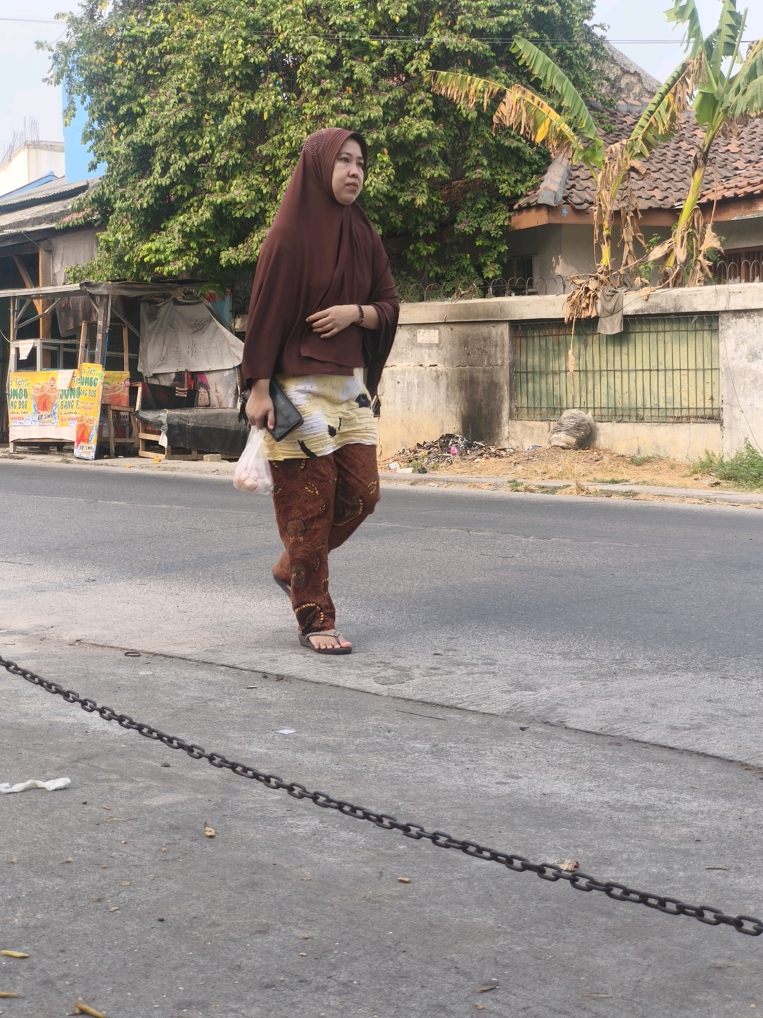 A woman wearing a brown hijab, a patterned long-sleeved top, and patterned pants walks along a paved road in an urban setting. She carries a black clutch and a plastic bag filled with items in her right hand. In the background, there are several structures, including a stall with colorful advertisements, a house with a tiled roof, and a wall with a metal fence. The area appears to be a street in Bekasi, Indonesia, with some signs of commercial activity and residential buildings. The road is lined with a concrete sidewalk and a metal chain barrier. There is also some debris and vegetation visible along the roadside. The lighting suggests it is daytime, with sunlight casting shadows.