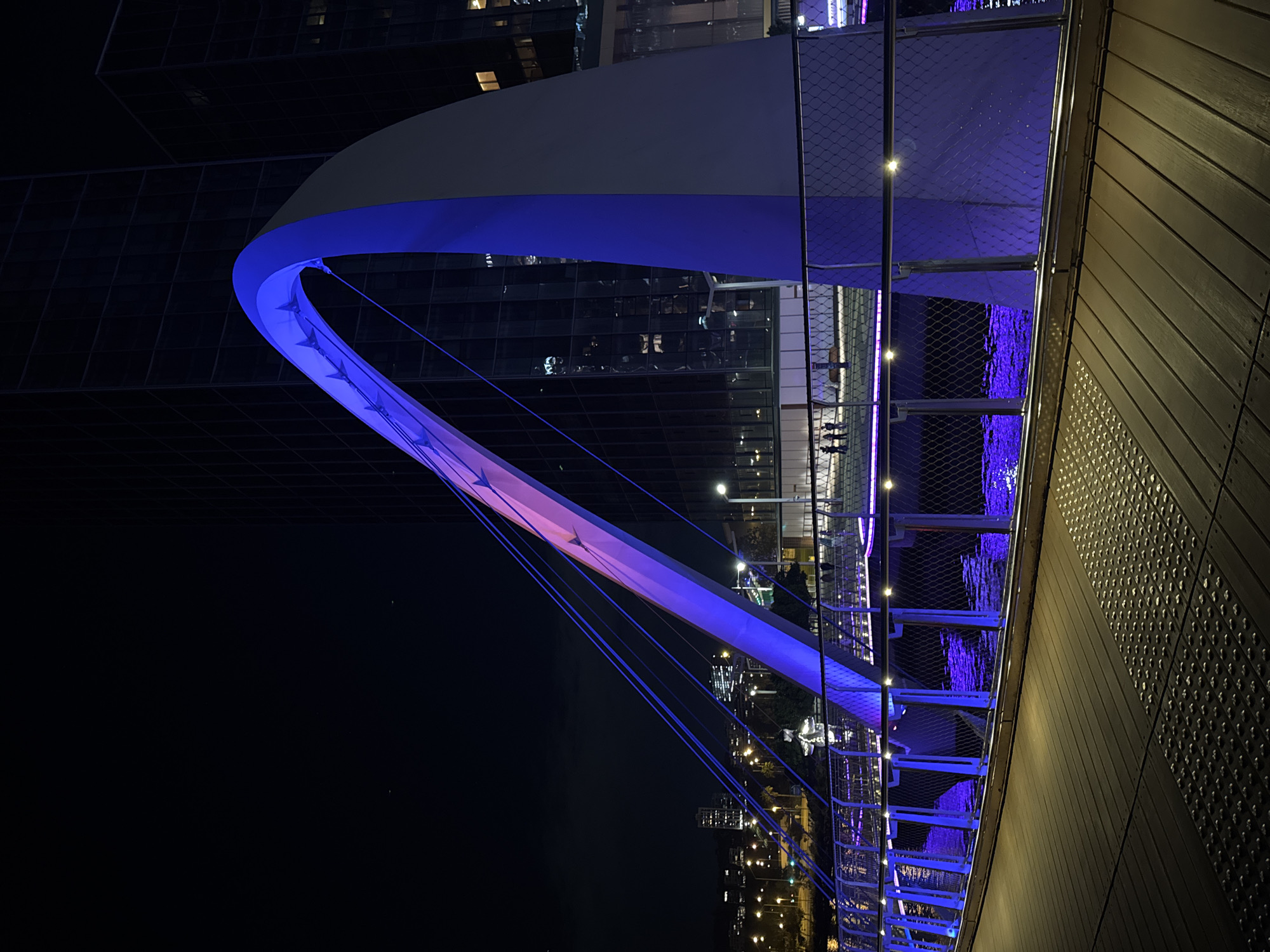 The image captures a vibrant night scene in Perth, Australia, dominated by a modern pedestrian bridge and its reflections on a body of water.
The main subject is a distinctly designed arch bridge, prominently illuminated with LED lights. The primary arch structure glows in a striking blue, transitioning to a gradient of pink and purple hues on the section further away from the viewer. The bridge's walkway features a mesh safety railing, along which numerous small, bright lights are embedded, casting a blue light downwards onto the water. Several cables are visible, connecting the bridge deck to the overhead arch.
On the right side of the frame, a wooden-clad structure with horizontal planks and a textured wall with a grid pattern of circular indentations forms part of a walkway or building adjacent to the bridge. Small, bright lights also line the edge of this structure.
Below the bridge, a body of water, likely a river or bay, reflects the vivid blue and purple lights of the bridge, as well as the scattered, warm-toned lights from buildings in the distant cityscape. Ripples on the water surface create dynamic, distorted reflections.
In the background, dark, modern buildings with illuminated windows are visible, typical of an urban environment at night. A few small figures can be seen walking on the bridge walkway, indicating human activity. The overall time of day is clearly night, characterized by the dark sky and the reliance on artificial lighting to define the scene. The weather appears clear. No visible text is discernible in the image.