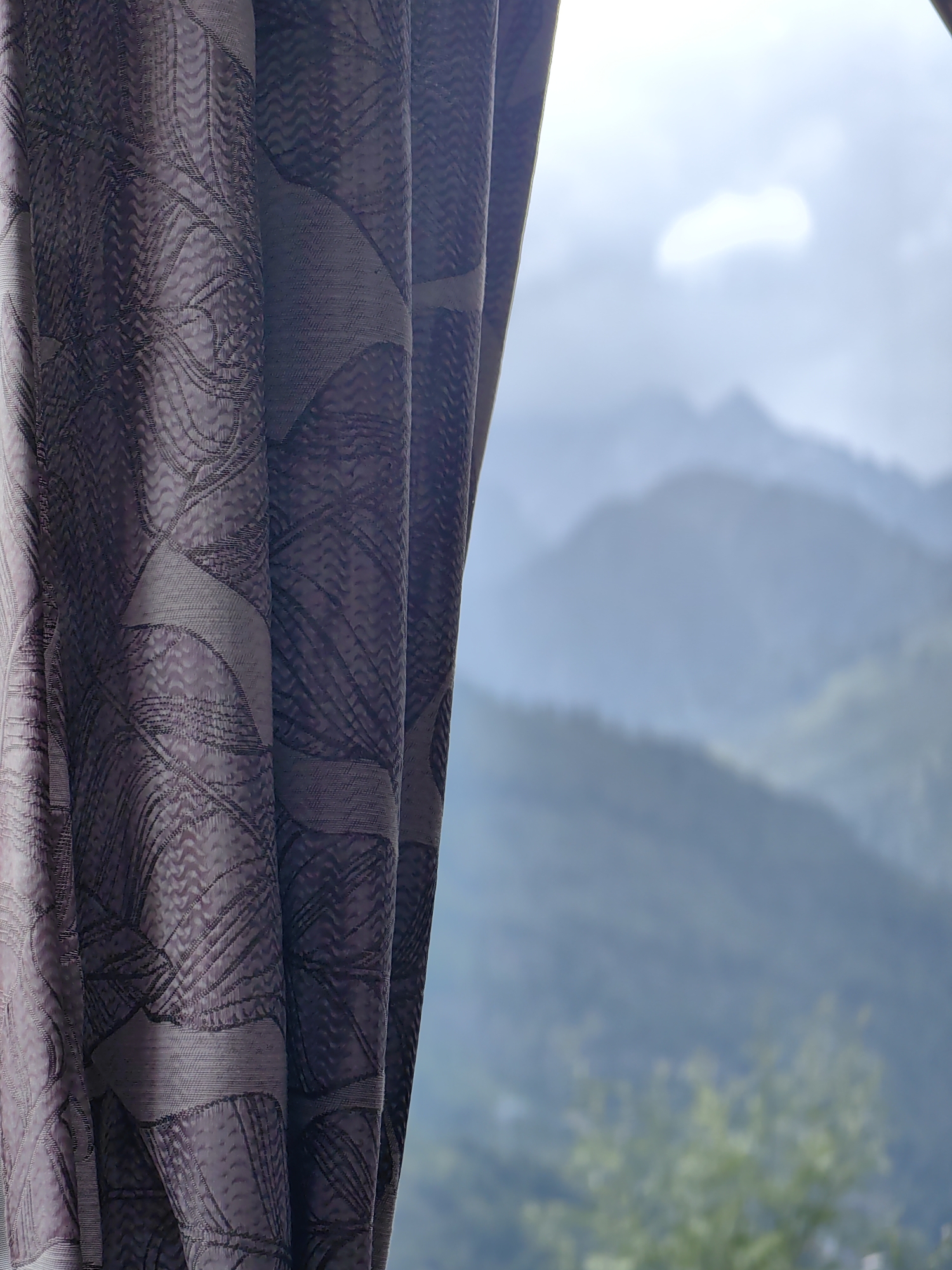 The image shows a close-up of a patterned purple curtain on the left side, with a blurred view of misty mountains in the background. The curtain has a geometric leaf-like pattern. Through a gap in the curtain, layers of green and blue mountains are visible, fading into the distance under a bright, overcast sky. The scene is suggestive of a view from a window or balcony in the Vashisht, India area, known for its scenic mountain landscapes.