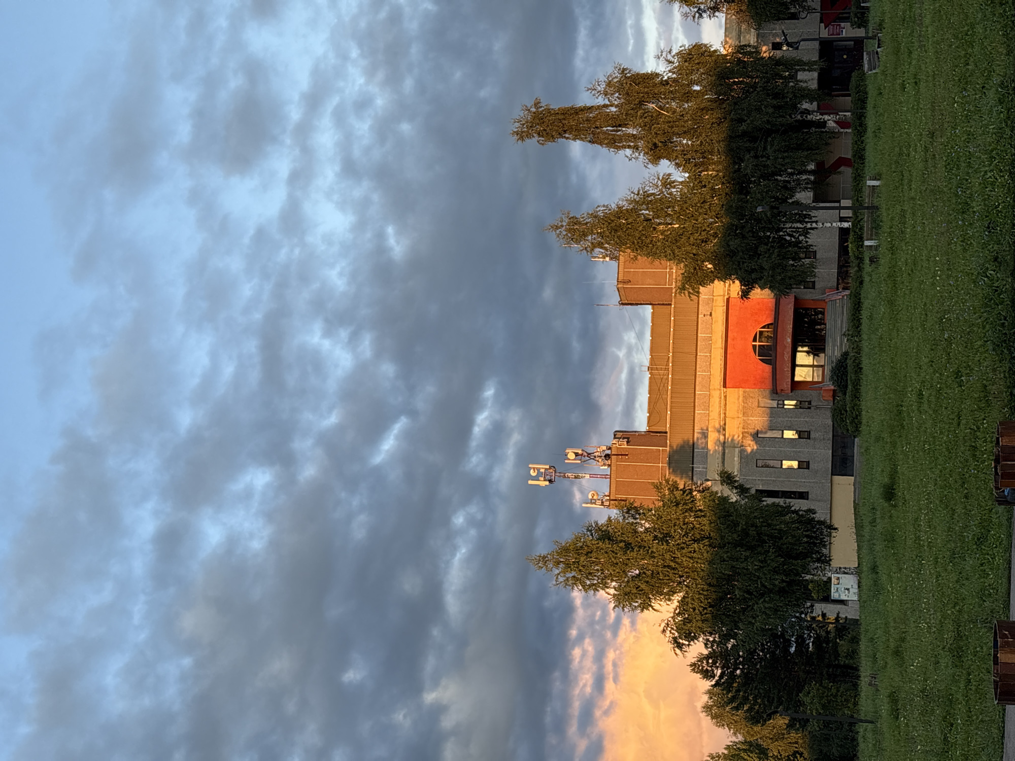 In the city of Markovskiy, Russia, a large, multi-story building with a light-colored concrete facade and a reddish-orange entrance is visible under a dramatic sky. The building is flanked by several trees, including a prominent birch tree with weeping branches. The sky is filled with dark, textured clouds, with a warm, orange glow from the setting sun illuminating the clouds on the left side of the frame. Several cellular antennas are mounted on the rooftop of the building. A grassy lawn stretches in front of the building, with a few benches and what appear to be planters or trash receptacles visible at the bottom of the frame. The scene appears to be a public space, possibly a park or recreational area, adjacent to the building.
