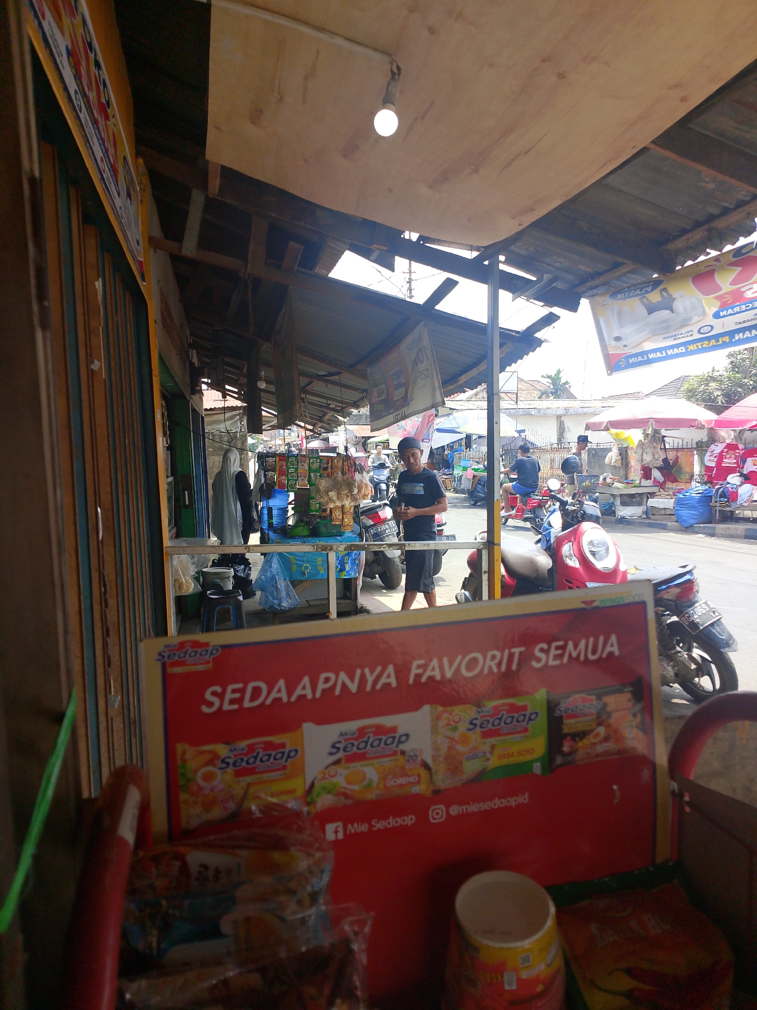 The image shows a street scene in Palembang, Indonesia, likely a market or roadside stall area. In the foreground, a red advertisement board is partially visible, displaying "SEDAPNYA FAVORIT SEMUA" along with images of instant noodle packets. To the left of the board, a red pole is visible, and behind it, a green object and what appears to be the edge of a shop with colorful vertical panels.
The background reveals a bustling street with several people and vehicles. A young man in a black t-shirt and a cap stands looking towards the left, with a motorcycle behind him. Further down the street, other individuals are seen, some on motorcycles, and various stalls with goods displayed under awnings and umbrellas. The architecture suggests a tropical climate, with open structures and corrugated metal roofing. The overall impression is a lively, everyday commercial setting in an urban Indonesian environment.