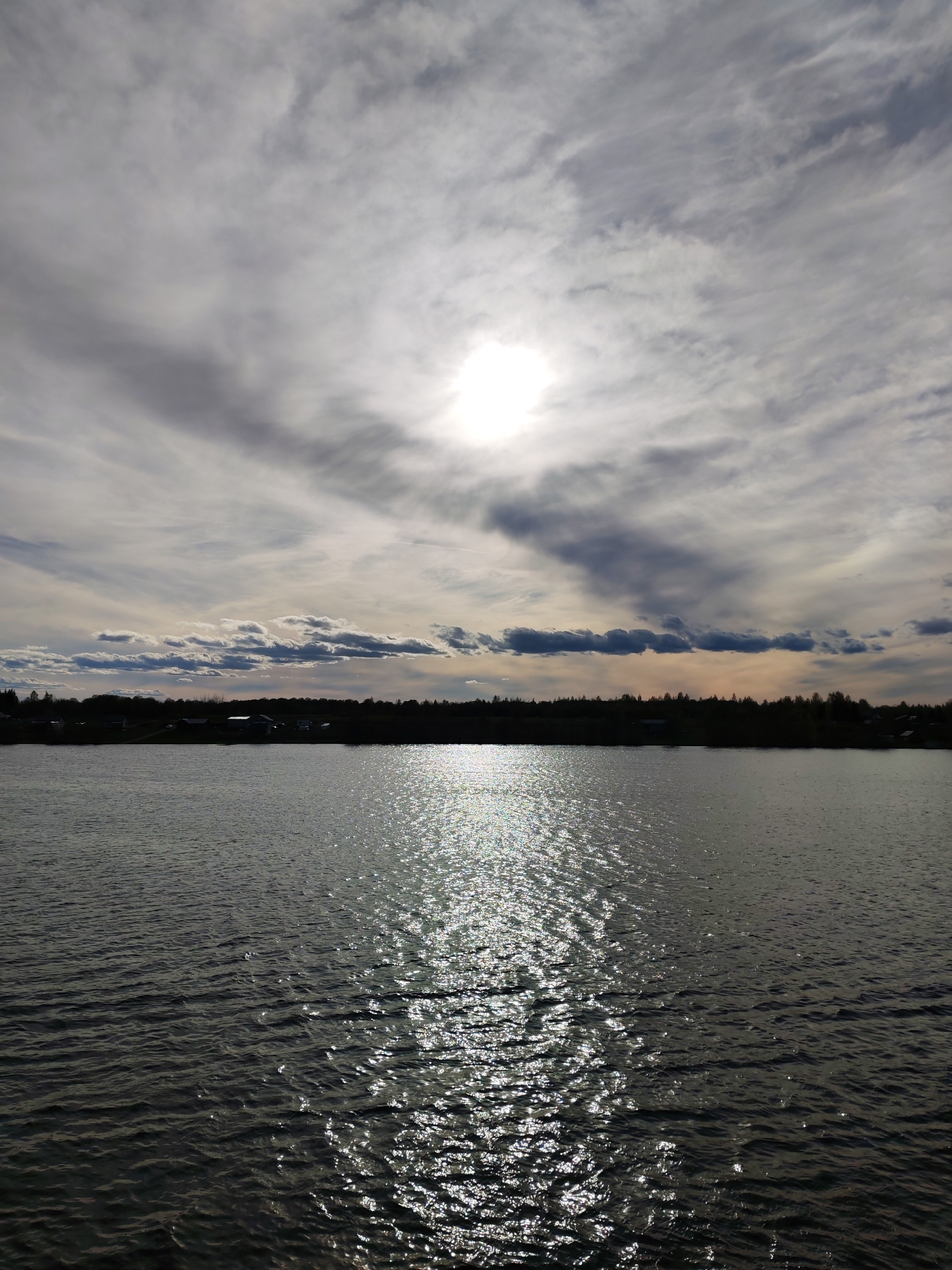 The image depicts a body of water, likely a lake or river, under a cloudy sky with the sun shining through. The water is reflecting the sunlight, creating a sparkling effect. In the distance, there is a treeline and what appears to be a settlement, possibly a village or town. Given the location, the scene is likely a rural or semi-rural area. Based on the provided information that the location is in the city of Раменье, Russia, it is reasonable to say that the image depicts nature in that city, perhaps on the outskirts of the main urban area. No people or animals are visible in the image.