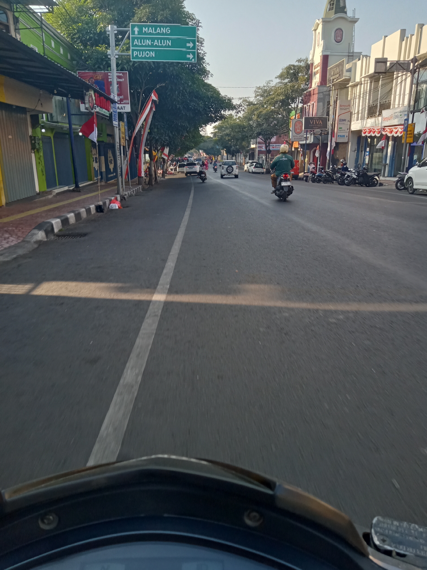 The image depicts a street scene in the city of Batu, Indonesia, likely along a main thoroughfare. A directional signpost indicates routes to "MALANG," "ALUN-ALUN," and "PUJON." On the left side of the road, there are several storefronts with closed metal shutters, some with awnings. The sidewalk is bordered by a curb. The road itself is paved with asphalt and has a white lane divider. Several vehicles are present, including motorcycles and cars. One motorcyclist, wearing a green jacket and helmet, is riding in the foreground of the traffic lane. Other motorcycles are parked along the right side of the road, in front of buildings that appear to be shops or businesses, one of which is identified as "VIVA BOUTIQUE." The buildings have multiple stories and display various signs. Trees line the street, providing some shade. The sky is clear and appears to be late afternoon, given the long shadows cast across the road. The perspective suggests the photo was taken from a moving vehicle, possibly a motorcycle.