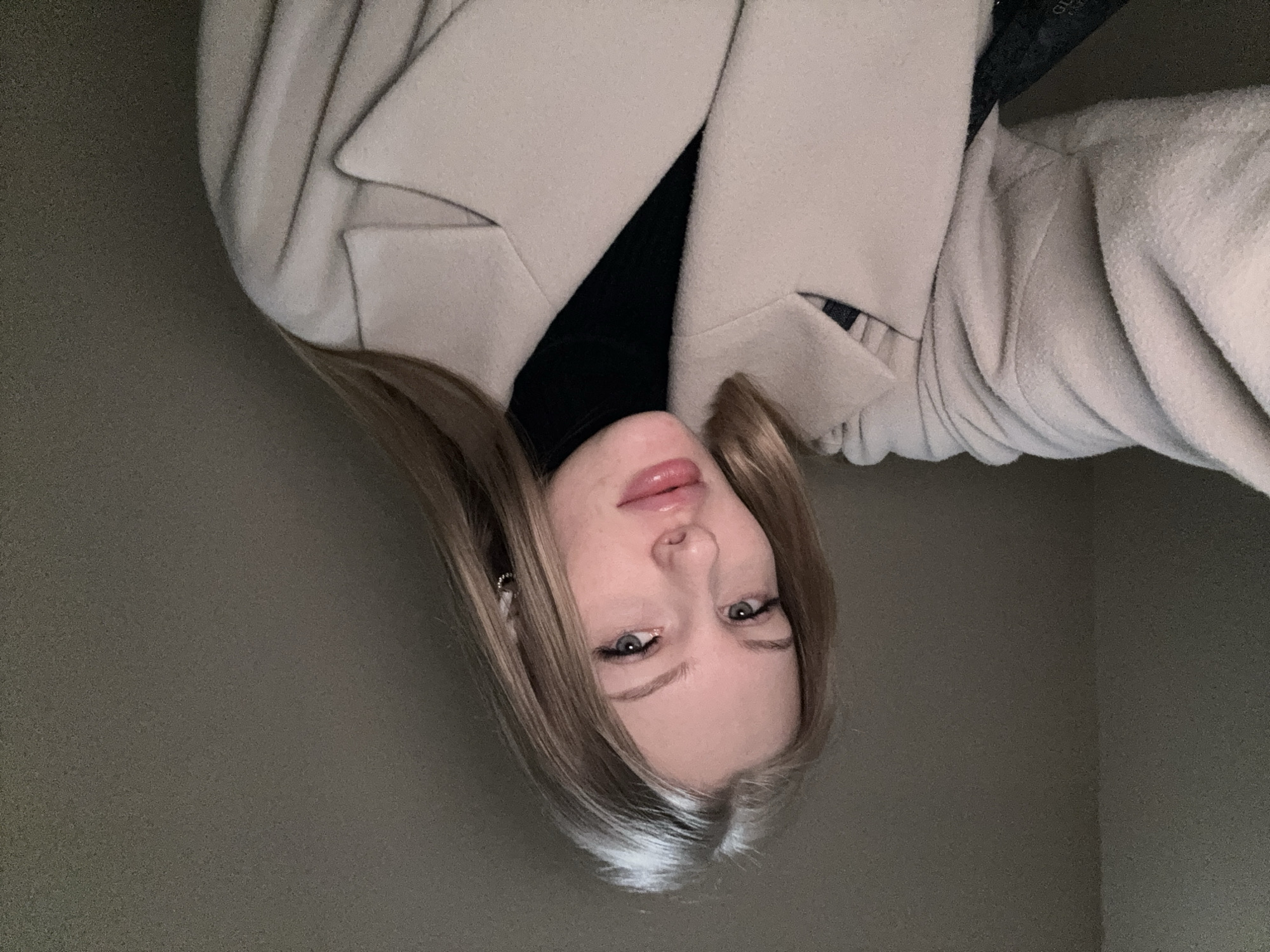 A photograph shows a person, presumably female, with long blonde hair. She is wearing a black turtleneck sweater and a light-colored overcoat. The setting appears to be indoors against a neutral wall. There's no clear indication of the location being Perm, Russia or any specific city.