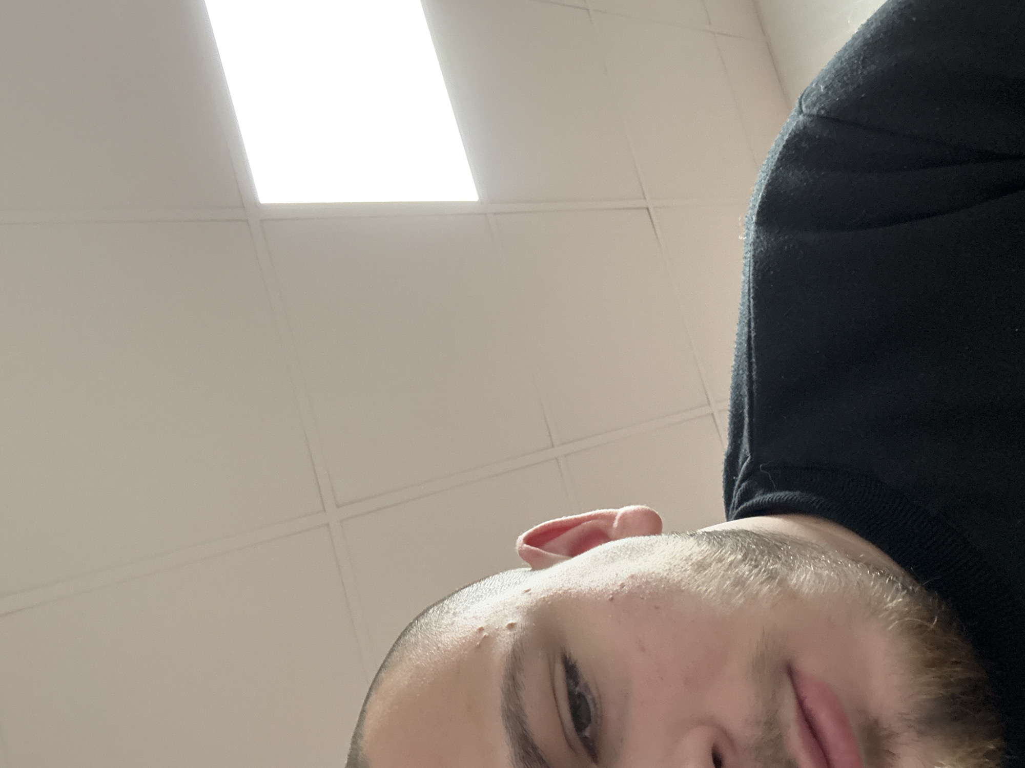 The image shows a low-angle, close-up view of a person's face, tilted upwards. The person has light skin, short hair, and a beard. An ear is visible near the top of the frame. The person is wearing a black top. In the background, there is a white ceiling with rectangular light fixtures. The scene appears to be indoors. There is no information available to determine if the location is in the city of Stara Zagora, Bulgaria.