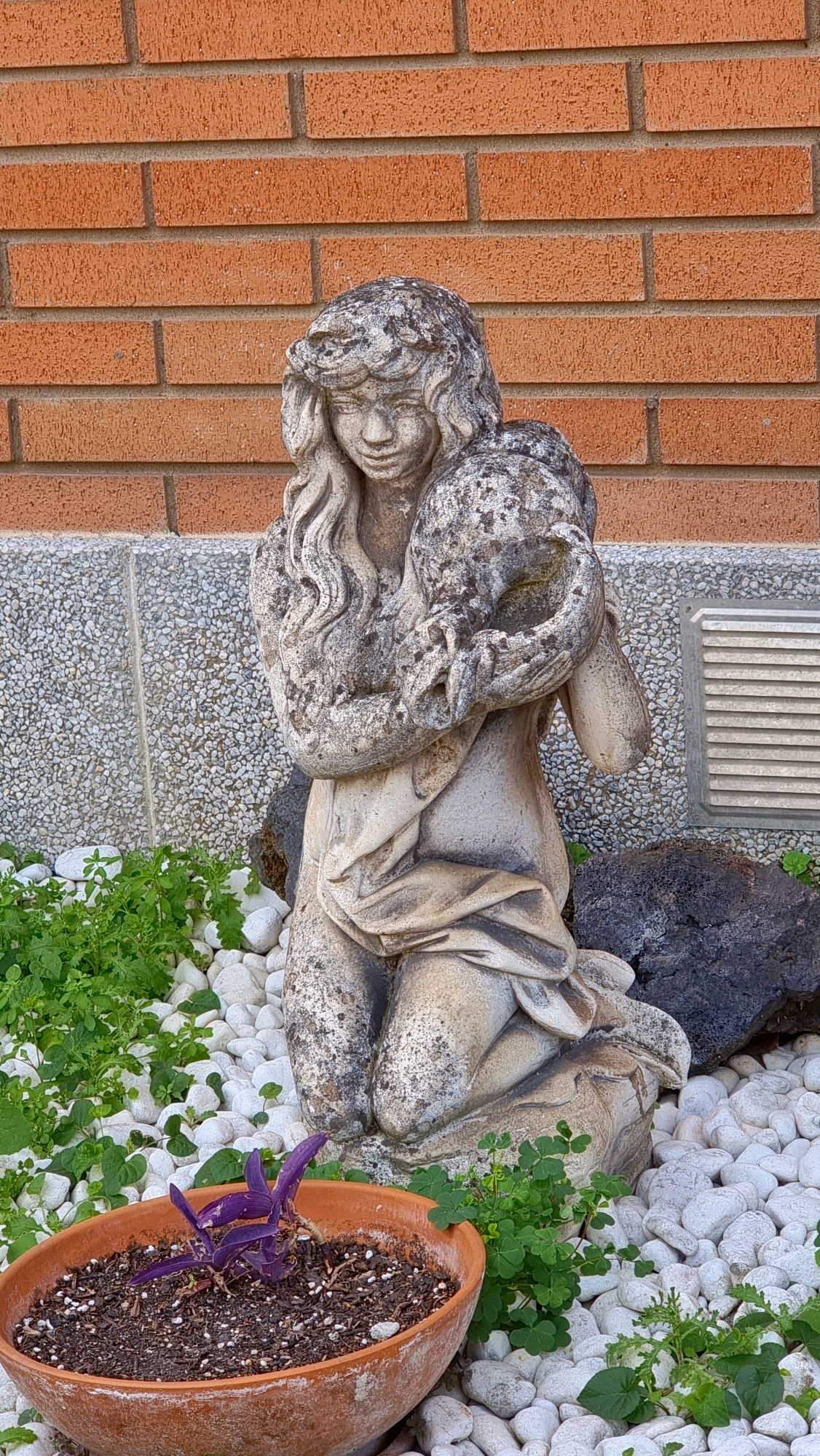 A weathered, light-colored stone statue of a kneeling woman is positioned in front of a red brick wall in La Canya, Spain. The statue depicts a woman with flowing hair, holding a spherical object in her arms. She is wearing a draped garment. The area around the statue is covered in white pebbles and small green plants. To the left of the statue, a terracotta pot contains dark soil and a small purple plant. In the background, a textured wall with a gridded vent is visible. The overall scene suggests an outdoor garden or decorative setting.