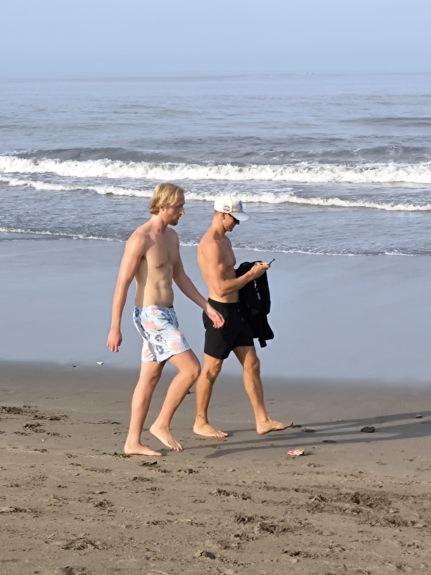 Two shirtless men walk along a sandy beach in Tibubeneng, Indonesia. The man on the left has blond hair and is wearing light blue patterned swim trunks. The man on the right wears a baseball cap and black swim trunks, holding a black garment and looking at a mobile phone. The beach is littered with debris. Waves are visible in the background.