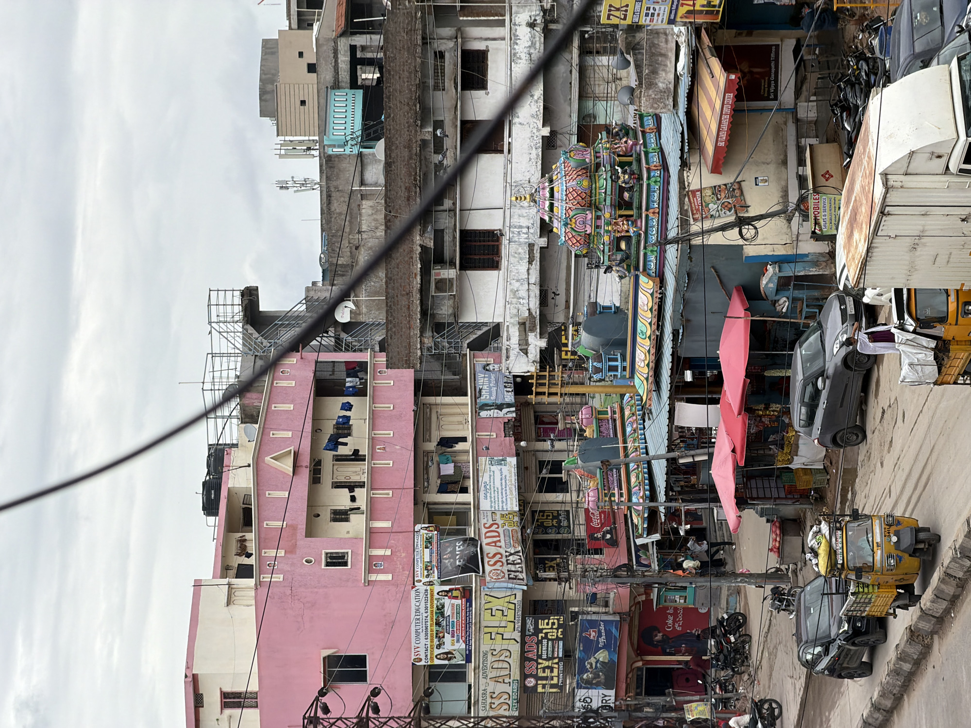 This is an eye-level, medium shot capturing a street scene in Shamshabad, India, under an overcast sky. The foreground is dominated by a yellow auto-rickshaw carrying goods, followed by a black SUV and a silver sedan. Beyond the vehicles, the street is lined with a mix of shops and multi-story buildings.
A prominent pink building on the left features multiple floors with windows and balconies, where laundry is hung out to dry. Various businesses are advertised on banners and signs attached to the buildings, including "SVV COMPUTER EDUCATION," "SAHASRA ADVERTISING," and "SS ADS FLEX." Further into the scene, a vibrant, colorful temple structure with intricate carvings and statues is visible. The temple has a tiered roof, painted in shades of pink, green, yellow, and blue, and is adorned with small figures of deities.
Adjacent to the temple are more shops with awnings, displaying goods for sale. Several motorcycles are parked along the roadside. People are seen walking on the street and in front of the shops. Utility poles and a dense network of electrical wires crisscross the view, indicating a busy urban environment. The buildings display a variety of architectural styles, from simple concrete structures to more ornate designs. The overall impression is of a bustling commercial area with a blend of traditional and modern elements.