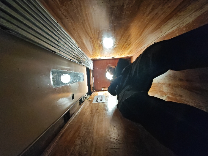 The image displays the interior of a narrow, enclosed wooden tunnel-like structure, likely a recreational slide, with the perspective directed upwards and outwards. This structure is potentially located within a public park or recreational area in Shumen, Bulgaria.
The interior surfaces of the tunnel – walls, floor, and ceiling – are constructed from light to medium-brown wood, exhibiting a worn, semi-glossy finish with visible grain and minor discolorations. On the left wall, a dark brown panel runs horizontally. Above this panel, a series of approximately nine parallel, horizontal metallic slats are affixed, secured by visible fasteners. Within the dark brown panel, a single, elongated oval-shaped opening with a reflective interior surface is present, appearing to be a window or port. Below this, smaller, indistinct rectangular apertures are visible along the wall's lower section.
At the distant end of the tunnel, positioned directly above the camera's viewpoint, one individual is partially visible. This person is looking downwards into the tunnel. The individual has short, light-colored hair and is wearing dark clothing, including what appears to be a jacket or hooded garment. Their face and immediate surroundings are illuminated by a circular, bright artificial light source positioned behind and above them, casting reflections on the wooden surfaces and the oval opening. A second, less intense light source is visible further in the background. On the wooden floor, directly beneath the individual's position, a small, flat white object, possibly a piece of paper or a mat, is discernable.