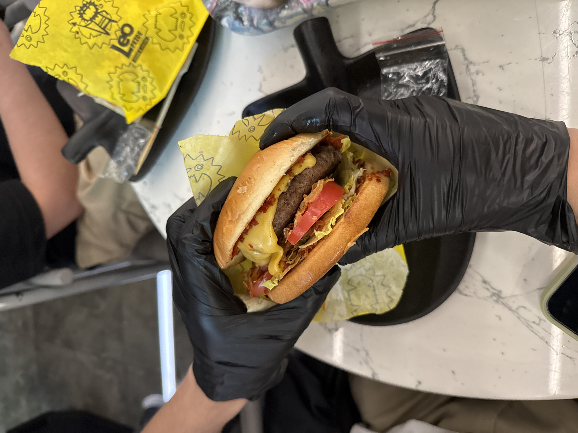 In Saint Petersburg, Russia, a close-up shot captures a person wearing black nitrile gloves holding a cheeseburger. The burger has a toasted bun, a beef patty, melted cheese, lettuce, and tomato. It is partially wrapped in yellow paper with a cartoonish sun graphic and the word "Leo" on it. The burger rests on a black plate, which is on a white marble-patterned table. To the right, a yellow paper bag with similar graphics and the text "Leo Moscow Pride" is visible, along with some plastic packaging. The background is blurred, showing hints of people and furniture, suggesting an indoor dining setting.