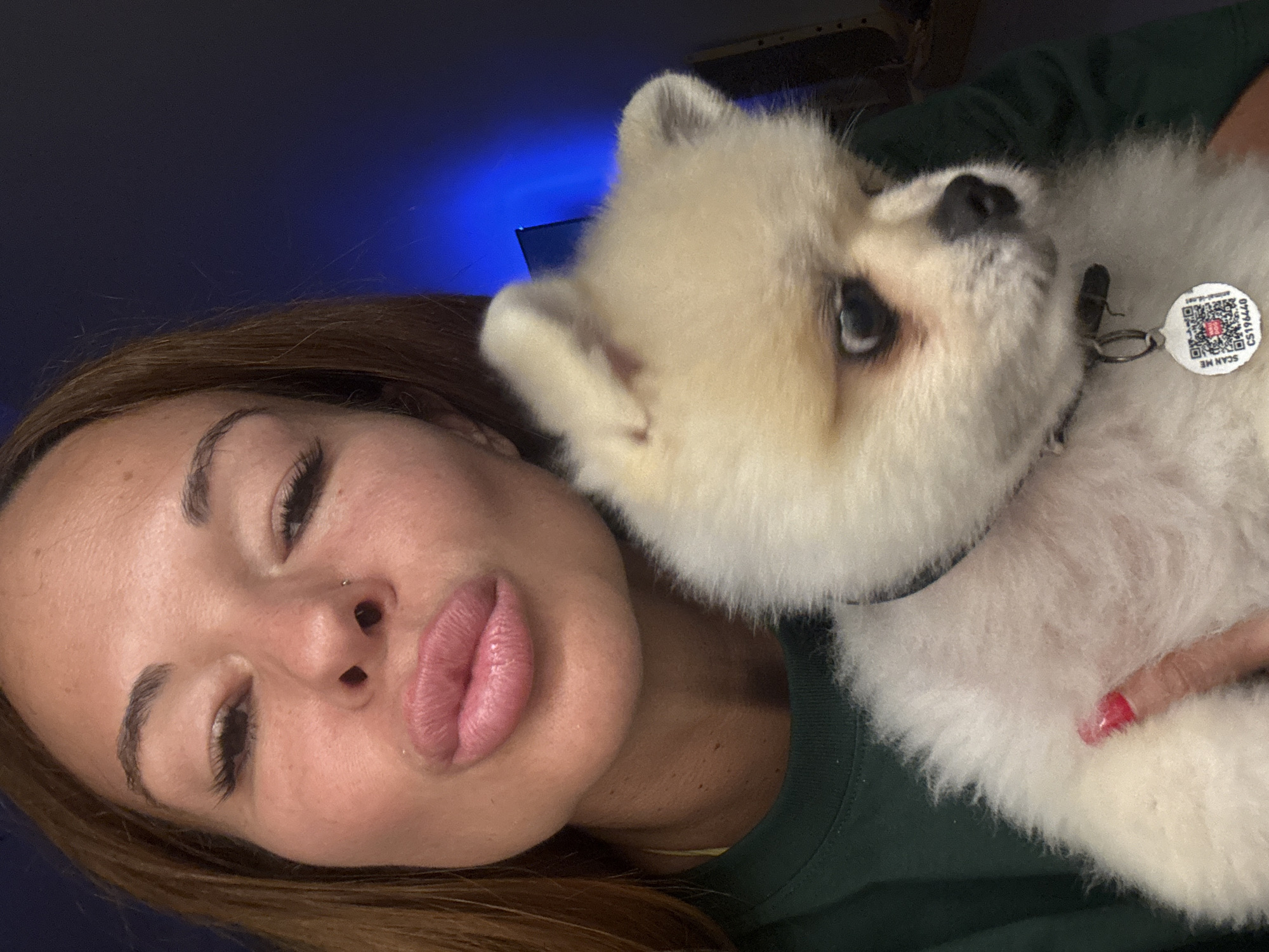 In this close-up, high-angle selfie, a woman and her fluffy white Pomeranian dog are the subjects. The woman, with her eyes closed and lips slightly parted, leans into her dog, who is nuzzling her cheek. She has long brown hair, full lips, and appears to be wearing makeup, including noticeable eyelashes and eyebrows. A small nose ring is visible. The dog has dark eyes and a black nose.
The background is dark, with a blurred blue light emanating from the top left, possibly from a screen or ambient lighting. The woman is wearing a dark green shirt, and the dog has a collar with a white tag featuring a QR code and the text "SCAN ME" and "animal-id.com" along with "CS196440". A woman's hand with red nail polish is visible on the dog's fluffy chest.
The overall impression is one of affection and intimacy between the woman and her pet. The lighting suggests it could be evening or indoors. No explicit weather or specific location cues beyond the provided context are visible in the image itself.
