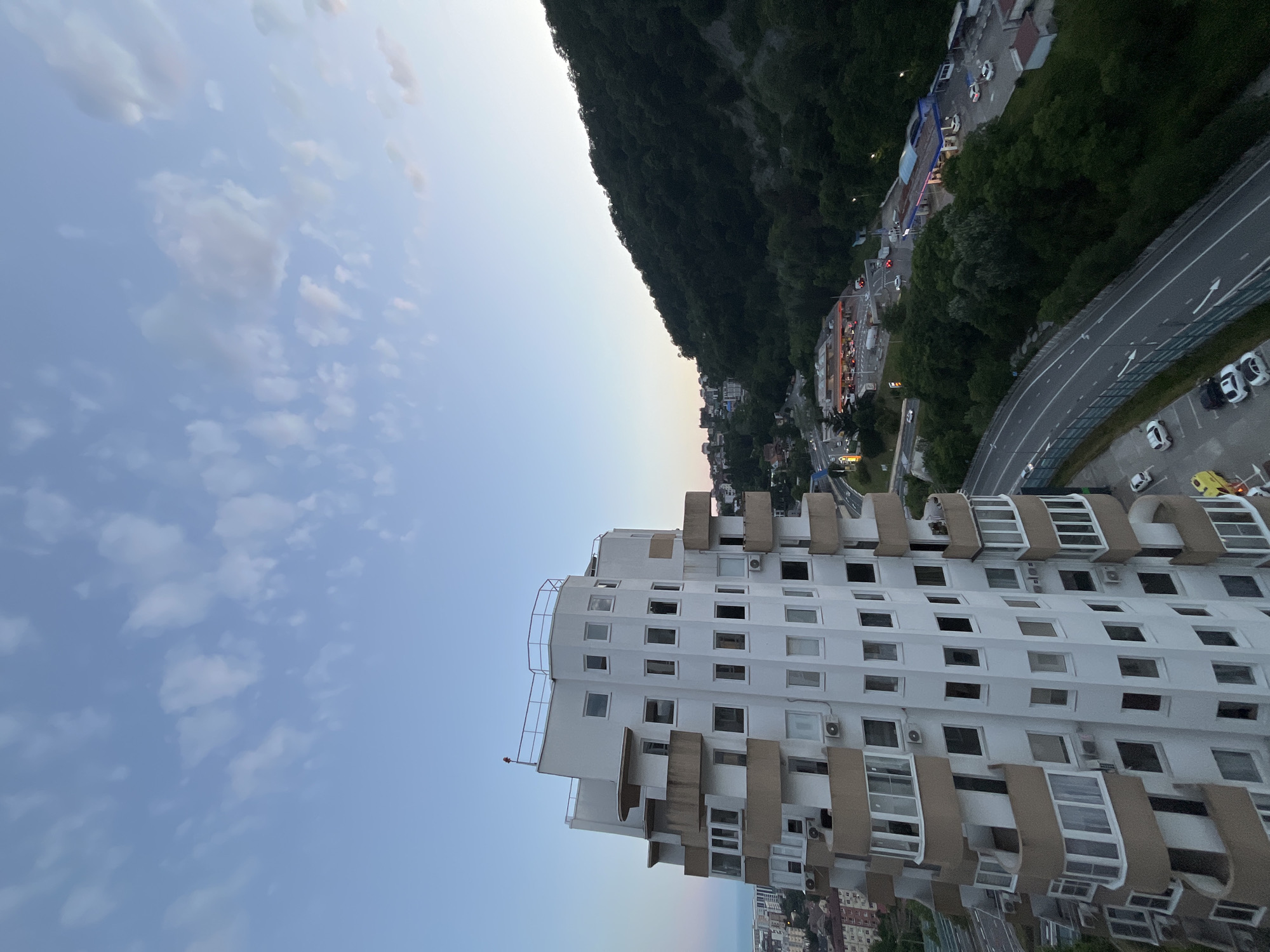 The image captures a high-angle view of a city scene in Sochi, Russia, at dusk. In the foreground, a distinctive white, multi-story building with curved balconies and numerous windows dominates the left side. Its unconventional, stepped design suggests a modern architectural style.
To the right and in the background, a densely forested hillside slopes upwards, meeting a pale blue sky with scattered wispy clouds. A winding road with several cars is visible traversing the base of the hill. Further down the road, a gas station and other commercial buildings are illuminated, indicating a commercial area. A few buildings that appear to be residential are nestled at the foot of the hill. The overall atmosphere is serene, with the fading light casting a gentle glow over the landscape. There are no people visible in the image.