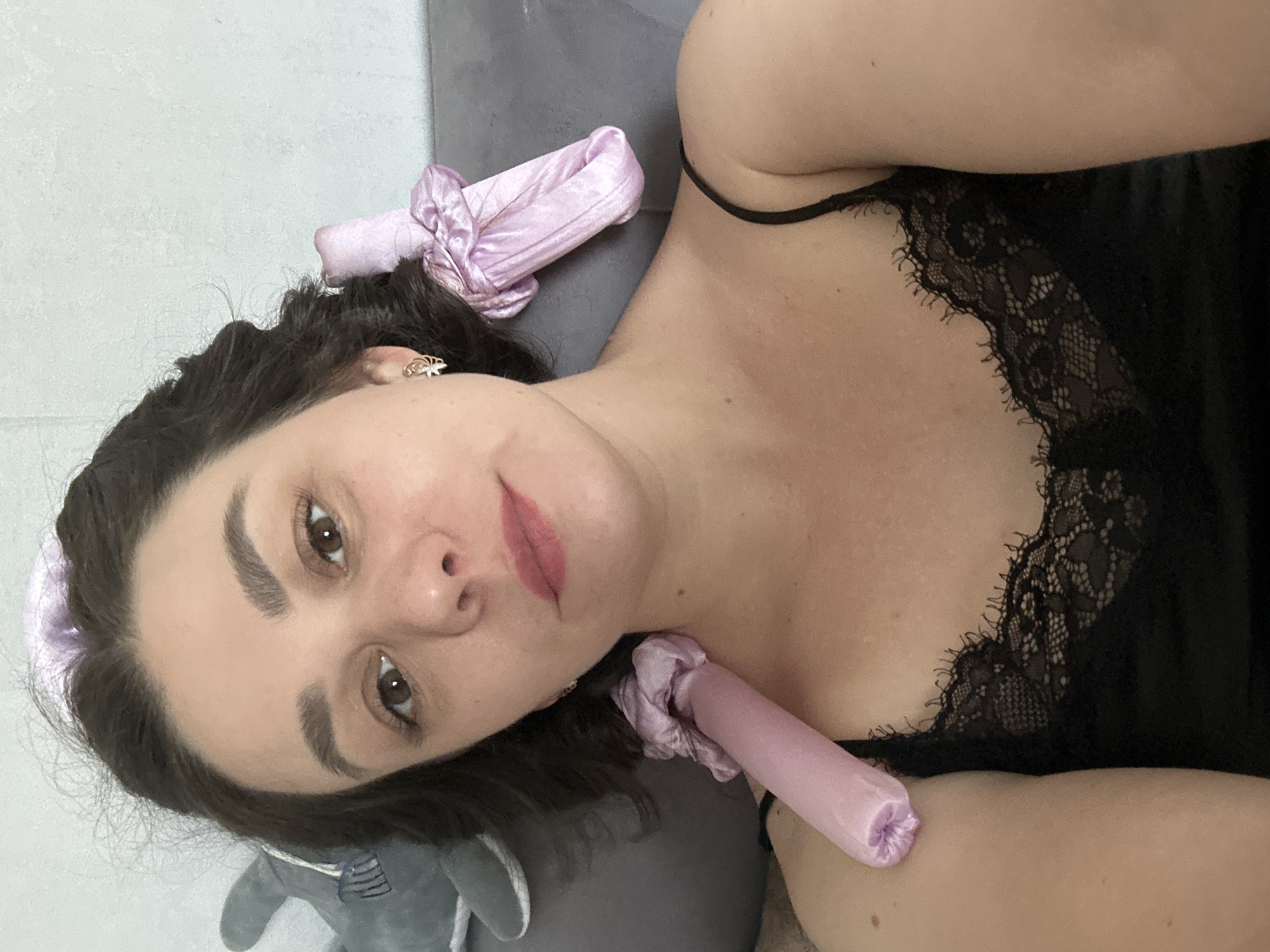 A woman lies on a gray surface wearing a black lace-trimmed camisole. Her dark hair is styled with two pink satin hair curlers, one on top of her head and another along her neck. She looks directly at the camera. A gray stuffed animal is visible in the lower left corner. The location is Chaikovsky, Russia.